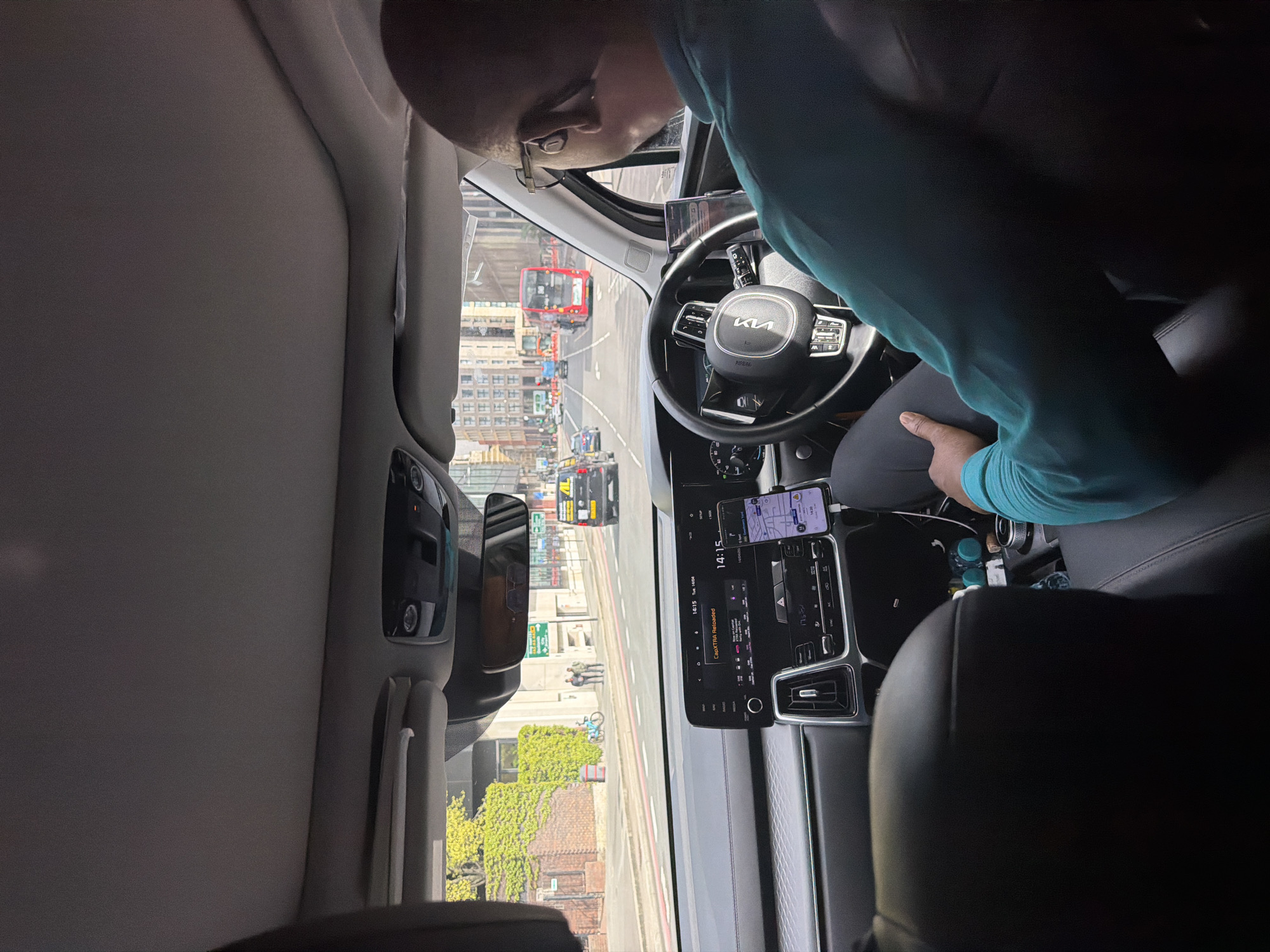 The interior of a Kia Sorento is presented from the passenger's point of view. The driver, a bald man with glasses and wearing a light blue shirt, is seen. The dashboard features a central display showing a navigation app and the time 14:14. A smartphone is mounted above the dashboard. The car's steering wheel has the Kia logo. Outside the vehicle, a view of London includes buildings, a red double-decker bus, and other vehicles. A brick building with ivy covering its exterior is visible.