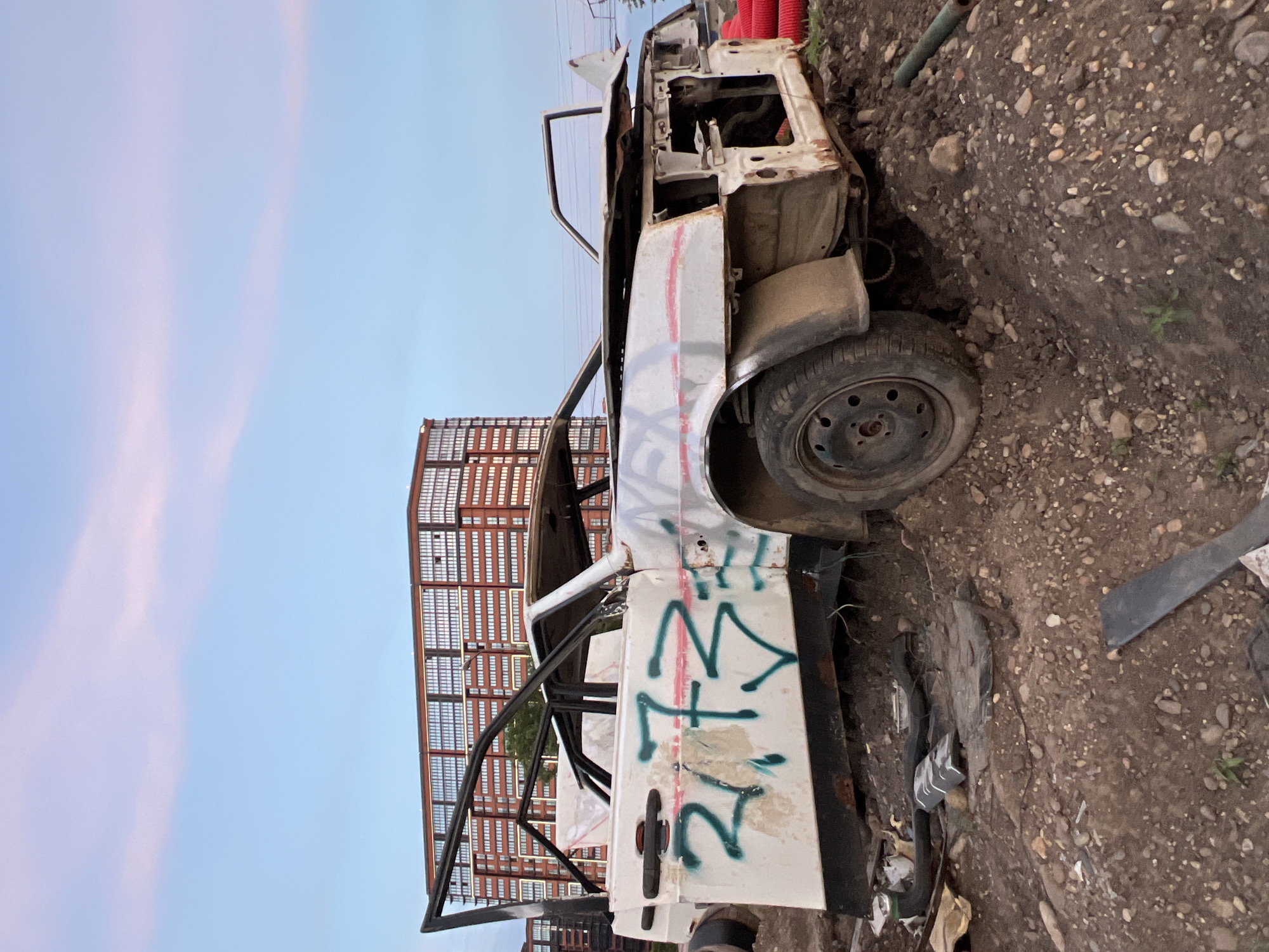 This is a photograph of a wrecked white car, possibly abandoned, lying on its side on a dirt and gravel lot. The car appears to be old and heavily damaged, with rust visible on its body. It has graffiti spray-painted on its doors, with green and red markings. The doors are open, and the interior is exposed, showing the car's stripped-down state.
In the background, a large, multi-story building with a red grid-like facade is visible, suggesting an industrial or urban setting. The sky is a soft blue with wispy pink clouds, indicating it might be early morning or late evening. The overall impression is one of neglect and decay, possibly in an area designated for vehicle disposal or in a derelict part of the city. There is no clear indication of people or any specific activity taking place, other than the implied abandonment of the vehicle. The ground is covered in small stones and dirt, with some sparse green vegetation growing in patches. The orientation of the image is rotated 90 degrees counter-clockwise.