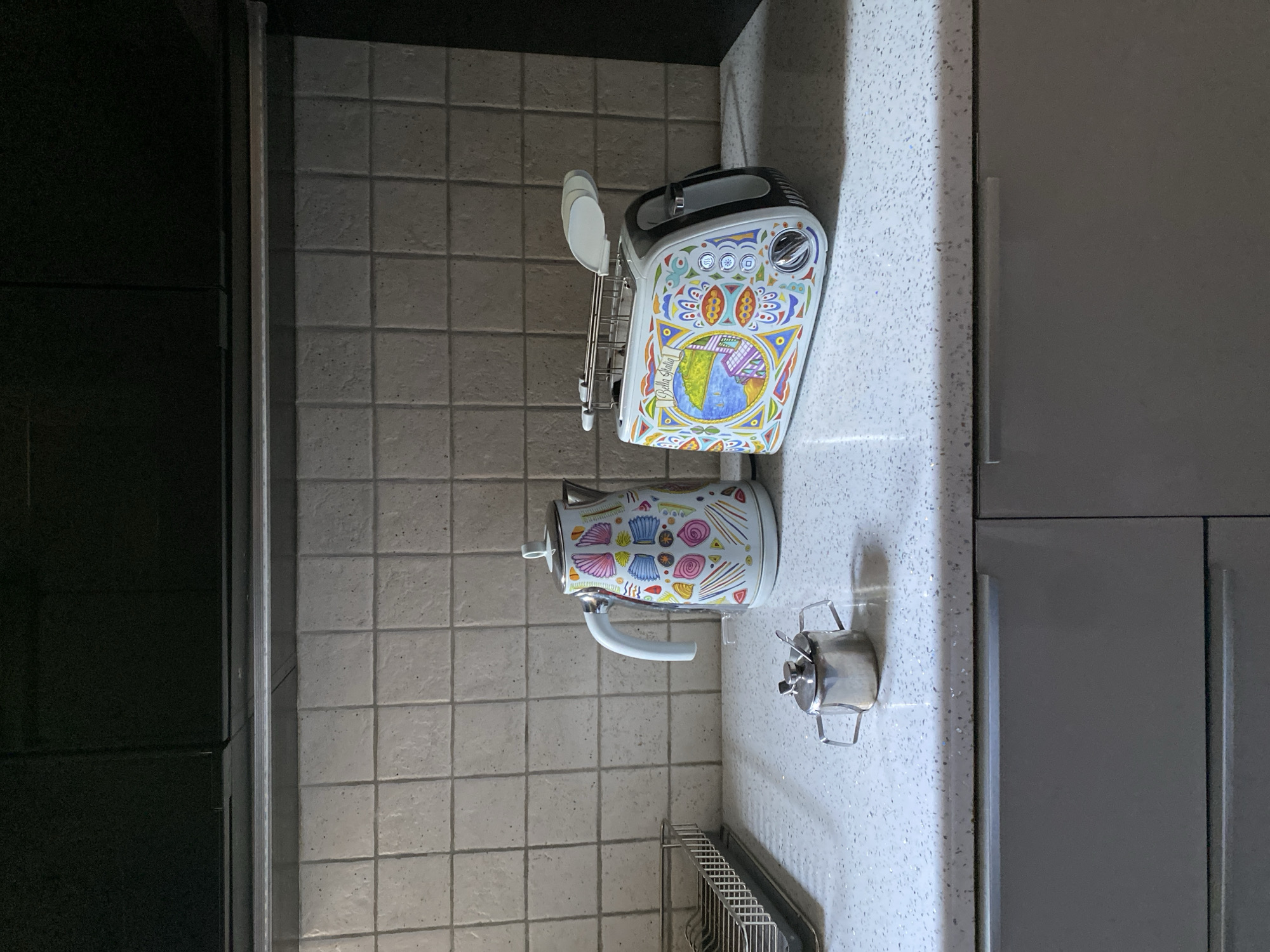 The image shows a kitchen counter in Wrocław, Poland, with a white kettle and a toaster decorated with colorful patterns. A small stainless steel sugar bowl with a spoon is placed in front of the kettle. The background features a beige tiled wall and dark cabinets. The counter is made of speckled white material, and a gray drawer is visible below. A stainless steel dish rack is partially visible on the left side of the image.