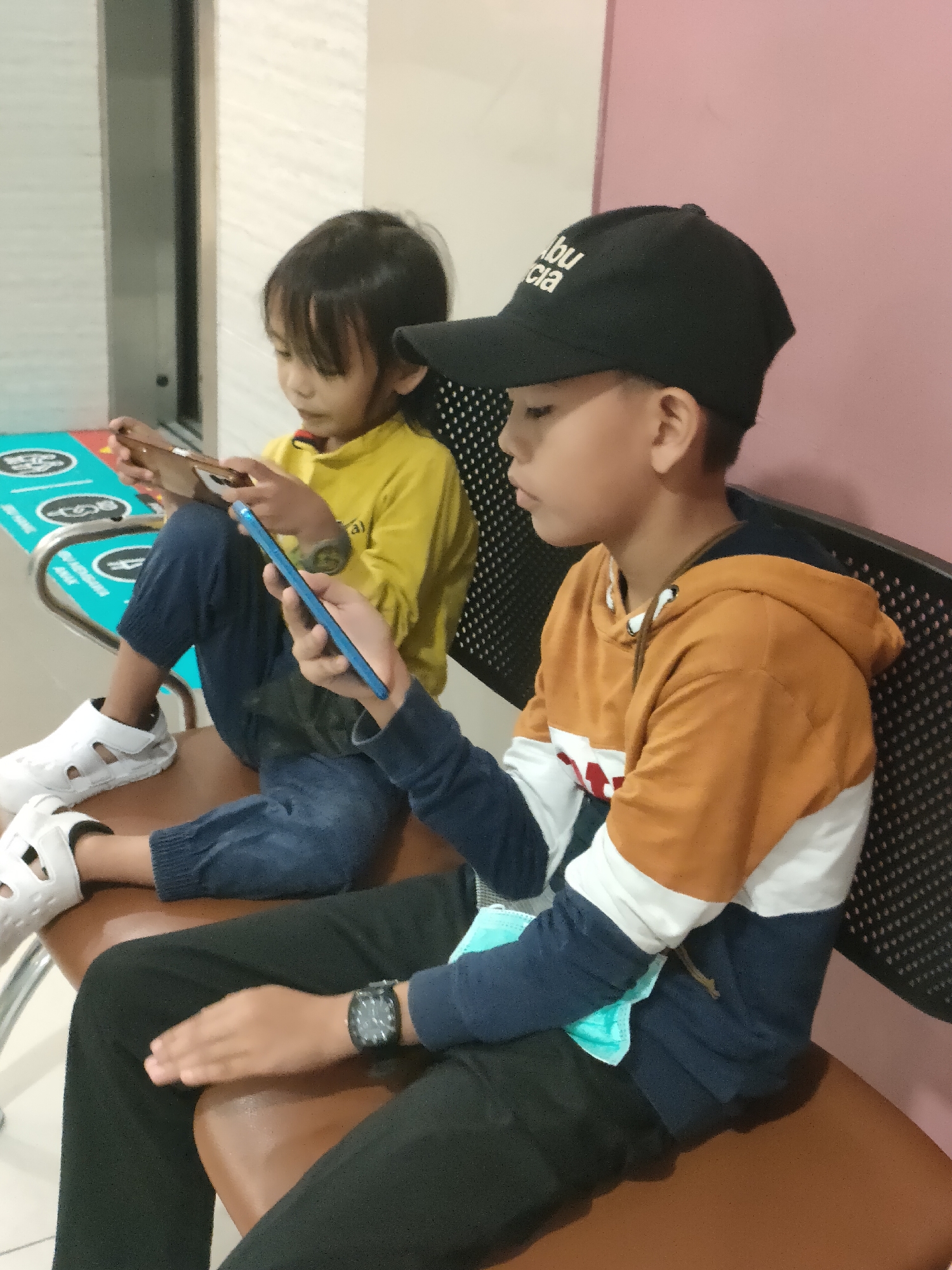 Two children, a younger girl and an older boy, are seated on a brown bench in what appears to be a waiting area. The younger girl, on the left, wears a yellow long-sleeved shirt and dark blue pants, with white sandals. She is looking down at a smartphone she holds in her hands. The older boy, on the right, wears a black baseball cap and a multi-colored hooded sweatshirt in shades of orange, white, and navy blue. He is also looking down at a smartphone, which is a vibrant blue color. He has a dark-colored wristwatch on his left wrist and a light blue disposable face mask hanging from his right sleeve. A textured, dark chair with a patterned back is behind them. To the left of the bench, a blue and teal-colored floor decal with circular graphics is visible. The background consists of a light-colored wall on the left and a light pink wall on the right. The overall setting suggests a public indoor space, potentially a clinic or office in Pekanbaru, Indonesia.