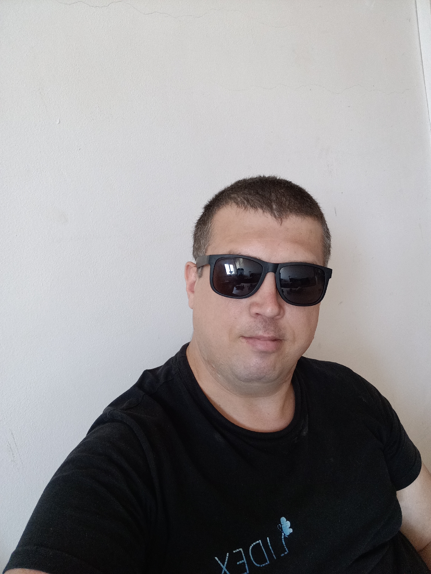 A medium shot captures a Caucasian man, appearing to be in his late 30s or early 40s, posing for a selfie against a plain, off-white wall. He has short, dark hair and is wearing black sunglasses that obscure his eyes. The man is dressed in a plain black t-shirt. The background wall has a slightly textured, uneven surface with a faint horizontal crack visible near the top. No specific landmarks or features indicate a location within Dalovy, Czechia. The lighting appears to be natural and diffuse, casting minimal shadows.