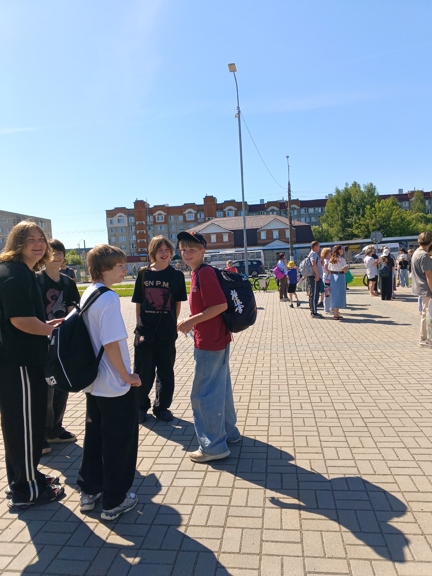 The image depicts an outdoor scene in Sudogda, Russia, under a clear blue sky on a sunny day. The foreground is a paved area constructed from light-colored rectangular interlocking pavers, displaying distinct, elongated shadows cast by people and structures, indicating a relatively low sun angle.
On the left side of the foreground, a group of six young males is present. From left to right:
1. A male with long, light brown hair, wearing a black t-shirt, black track pants with white side stripes, and white sneakers. He is smiling, looking towards the camera, and holding an indiscernible item in his left hand.
2. Partially obscured behind the first, a male with dark hair, wearing a dark t-shirt and dark pants.
3. A male wearing a white t-shirt, dark pants, and grey-and-white sneakers. He carries a black backpack and is smiling while looking towards the right.
4. A male with dark, medium-length hair, wearing a black t-shirt with white text ("EN P.M." and "NOWHERE AT I.AM." with a graphic), dark pants, and a black fanny pack. He is smiling and looking towards the camera.
5. A male with medium-length light brown hair, wearing a red t-shirt, light blue jeans, and light-colored sneakers. He carries a black backpack over his right shoulder and is smiling while looking towards the camera.
6. Another male is partially visible between the first and second, mostly obscured.
This group appears to be interacting and pausing on the paved surface.
In the mid-ground on the right, a larger group of approximately 15-20 individuals, including adults and some children, is gathered. They are standing or walking on a similar paved area, with some appearing to wait, possibly near a public transport stop where a white bus is partially visible behind them. A dark car is also visible further back.
The background features multiple multi-story residential buildings, primarily light-colored (beige/white) with reddish-brown roofs. A distinct two-story building with a reddish-brown tiled roof and light brown walls is visible in the center-right background, possibly a commercial or public establishment. Green foliage from trees is visible behind these structures on the right.
Two streetlights are visible: a tall metal pole with a rectangular light fixture on the left side of the frame, and a smaller, less distinct one further back. Utility lines traverse the upper mid-ground horizontally. The sky is clear and light blue.