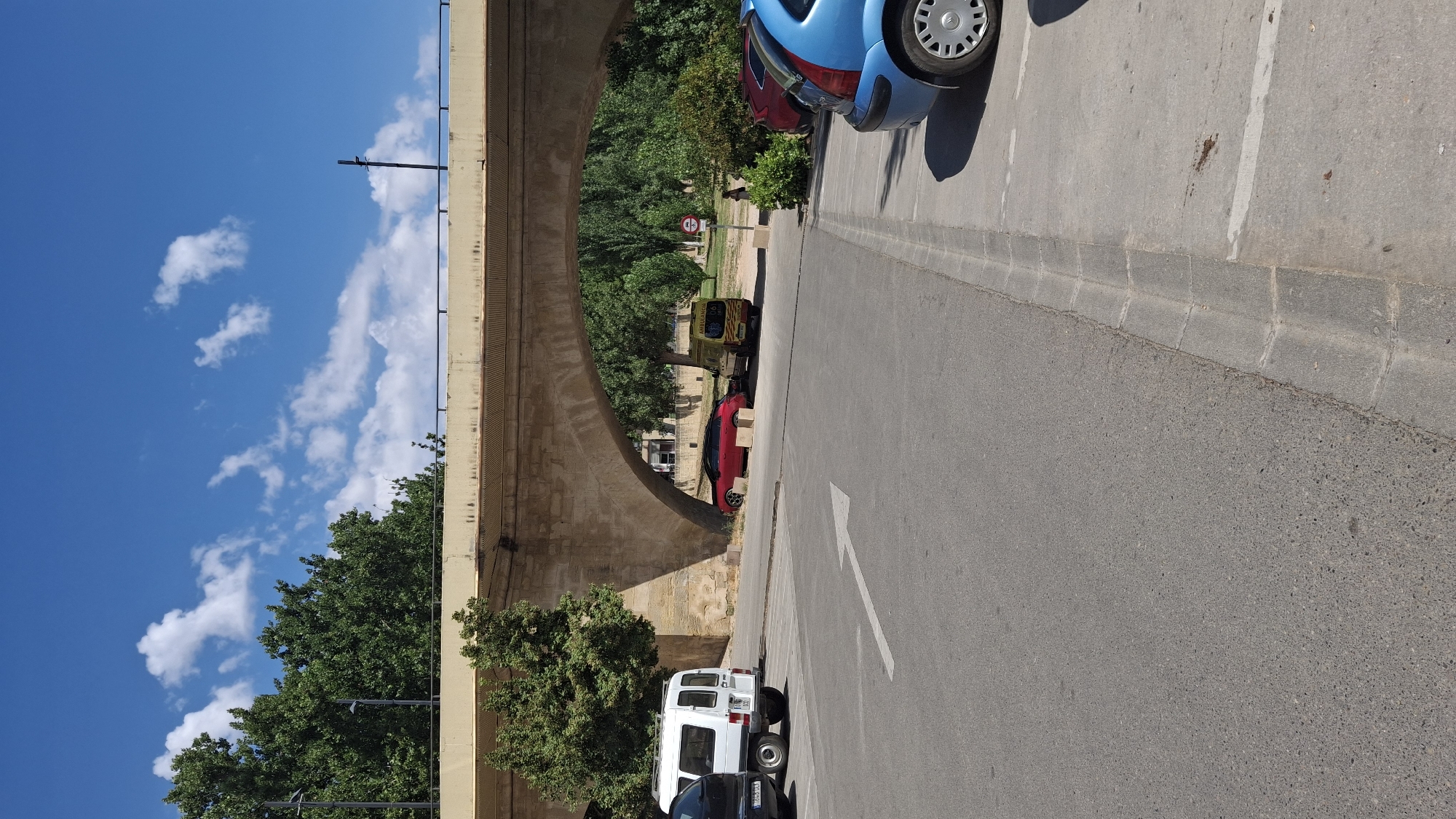 The image captures a scene beneath a concrete arch bridge, likely part of the Alcañiz ring road in Spain. A white van is parked on the left, followed by a red sports car. Further to the right, under the arch, is a yellow ambulance with the word "AMBULANCIA" visible on its side. Beyond the ambulance, there's a circular red and white sign, possibly indicating a speed limit or prohibition. A person is visible standing in the background near some green bushes. On the right side of the image, the rear of a blue car is parked, with its wheel and part of its body visible. The ground is paved with asphalt in the foreground, with a concrete curb separating it from a parking area. Lush green trees line the background, and the sky is blue with scattered white clouds. The overall impression is that of a sunny day in an urban or semi-urban environment.