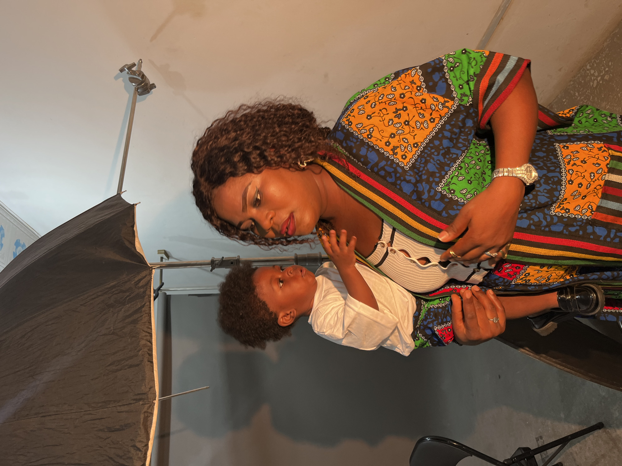 A dark-skinned woman in a vibrantly patterned, colorful dress is holding a young Black child. The woman has dark, curly hair and is wearing makeup, including red lipstick and eyeshadow. She has jewelry on her left hand, including rings and a watch. The child, wearing a white t-shirt, looks away from the camera. They are in an indoor setting that appears to be a studio, with a large black umbrella-like light diffuser in the background, a white wall, and some lighting equipment. The scene suggests a photo shoot is in progress or has recently concluded. The location is specified as Gura Top, Nigeria.