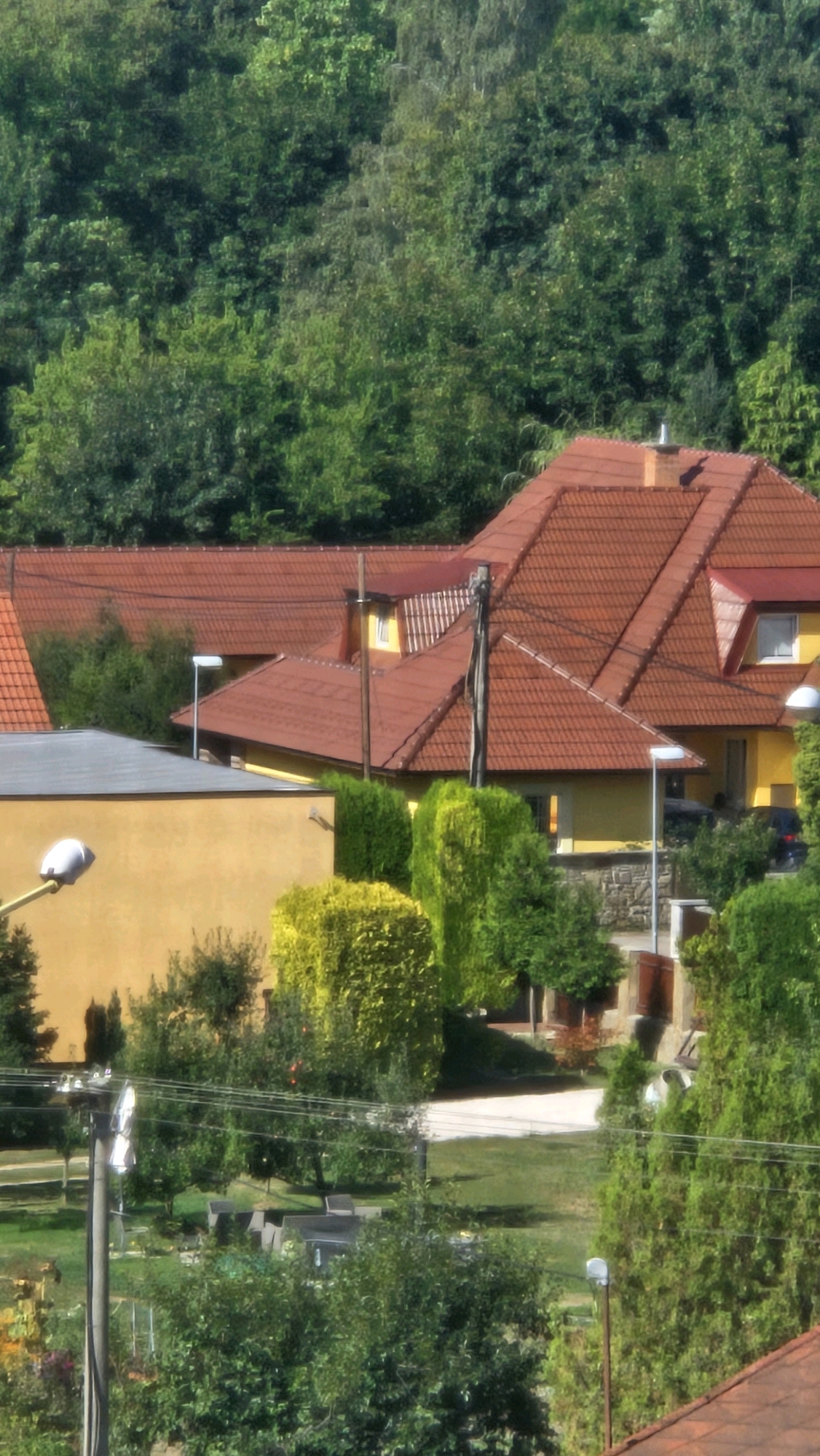 The image shows a residential area with several houses, characterized by their red tile roofs. The houses are nestled against a backdrop of lush green trees, suggesting a location with significant natural foliage. In the foreground, there are various bushes and trees, some of which are meticulously trimmed, indicating well-maintained gardens. A streetlamp is visible in the lower left corner, and power lines traverse the scene, suggesting the presence of infrastructure. The overall impression is a peaceful, semi-rural setting within a city. The presence of parked cars, visible in the background, further confirms it's a residential area. The architecture of the houses, particularly the roof designs and window placements, hints at a European style. The weather appears to be clear and sunny, based on the bright lighting and the vivid colors of the vegetation. It is plausible that this scene depicts a residential neighborhood in the city of Prešov, Slovakia, given the characteristic building styles and the abundance of greenery often found in such locales.