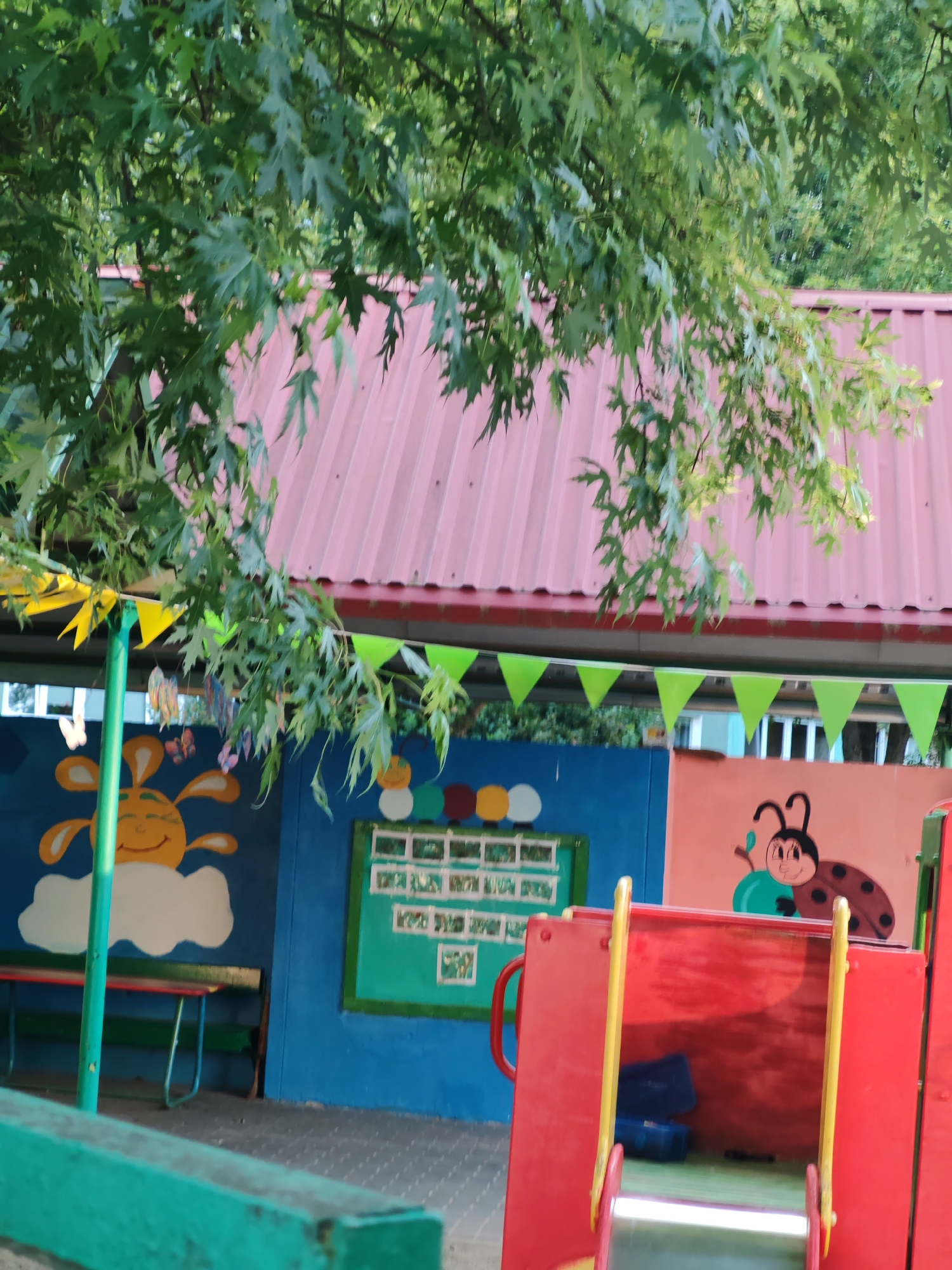 The image shows a brightly painted wall with cartoon characters, possibly in a playground or school setting. A red slide is visible in the foreground. The wall features a smiling sun character on a blue background to the left, and a ladybug character on a pink background to the right. Between them, a green board displays some colorful circles at the top and what appears to be a grid of photos or drawings below. Green and yellow pennant banners are strung along the top of the wall. The roof of a building, with a red, corrugated metal surface, is visible above the wall. Lush green foliage from trees hangs down over the scene, partially obscuring the roof and banners. A green bench with a red seat is positioned in front of the blue section of the wall. The ground is paved with interlocking stones. The overall scene suggests an outdoor recreational area designed for children.