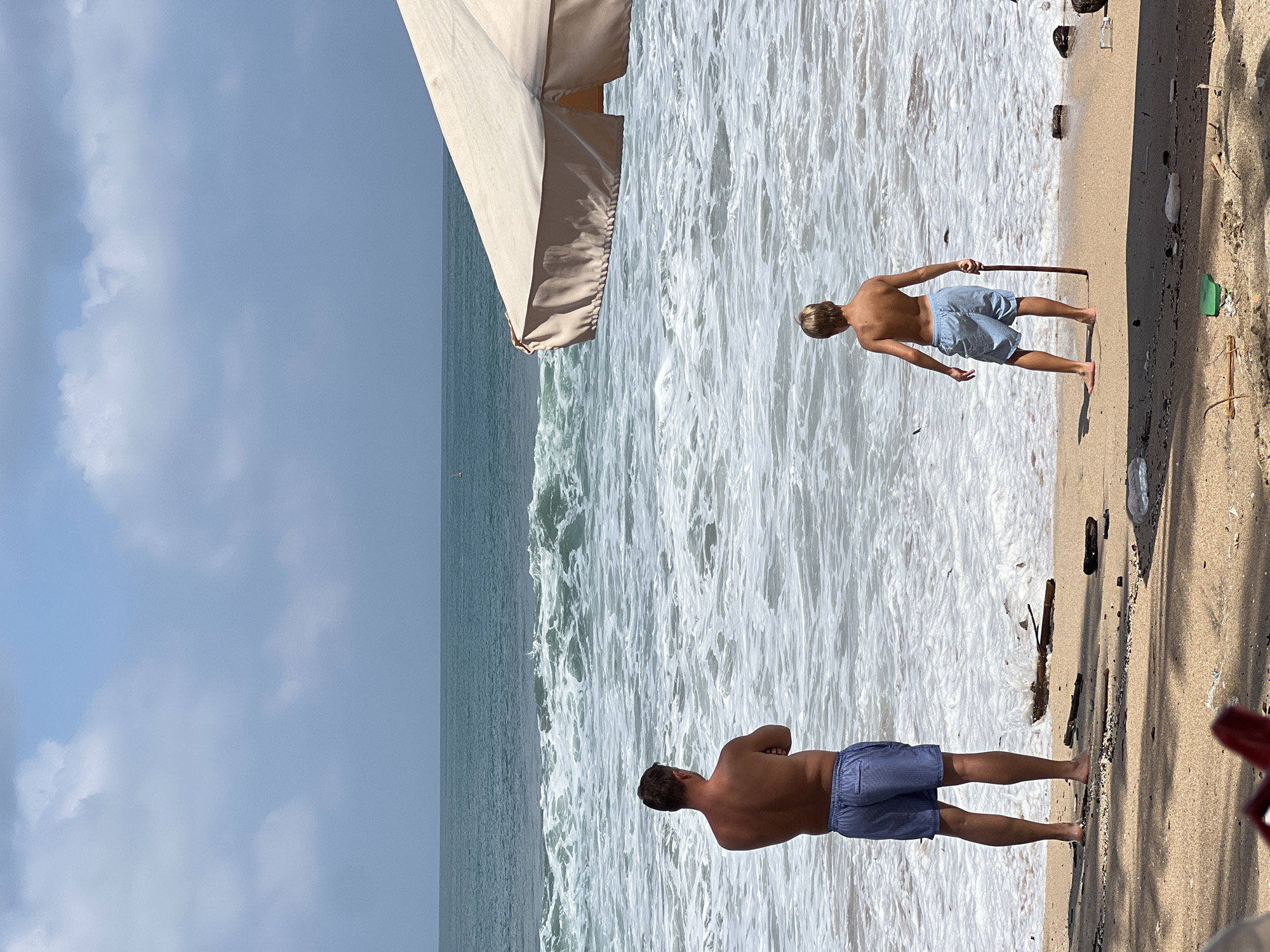 The image depicts a beach scene in Kuta, Indonesia, on a sunny day with scattered clouds. Two individuals, an adult male and a young boy, stand on the sandy shore facing the ocean. The man, wearing blue striped shorts, has his arms crossed and is looking out at the waves. The boy, also in blue shorts, holds a stick and faces the water. The sea is rough with white foam from the waves. A large beige beach umbrella is partially visible on the right side of the frame, casting a shadow on the sand. Debris, including wood and plastic, is scattered on the beach.