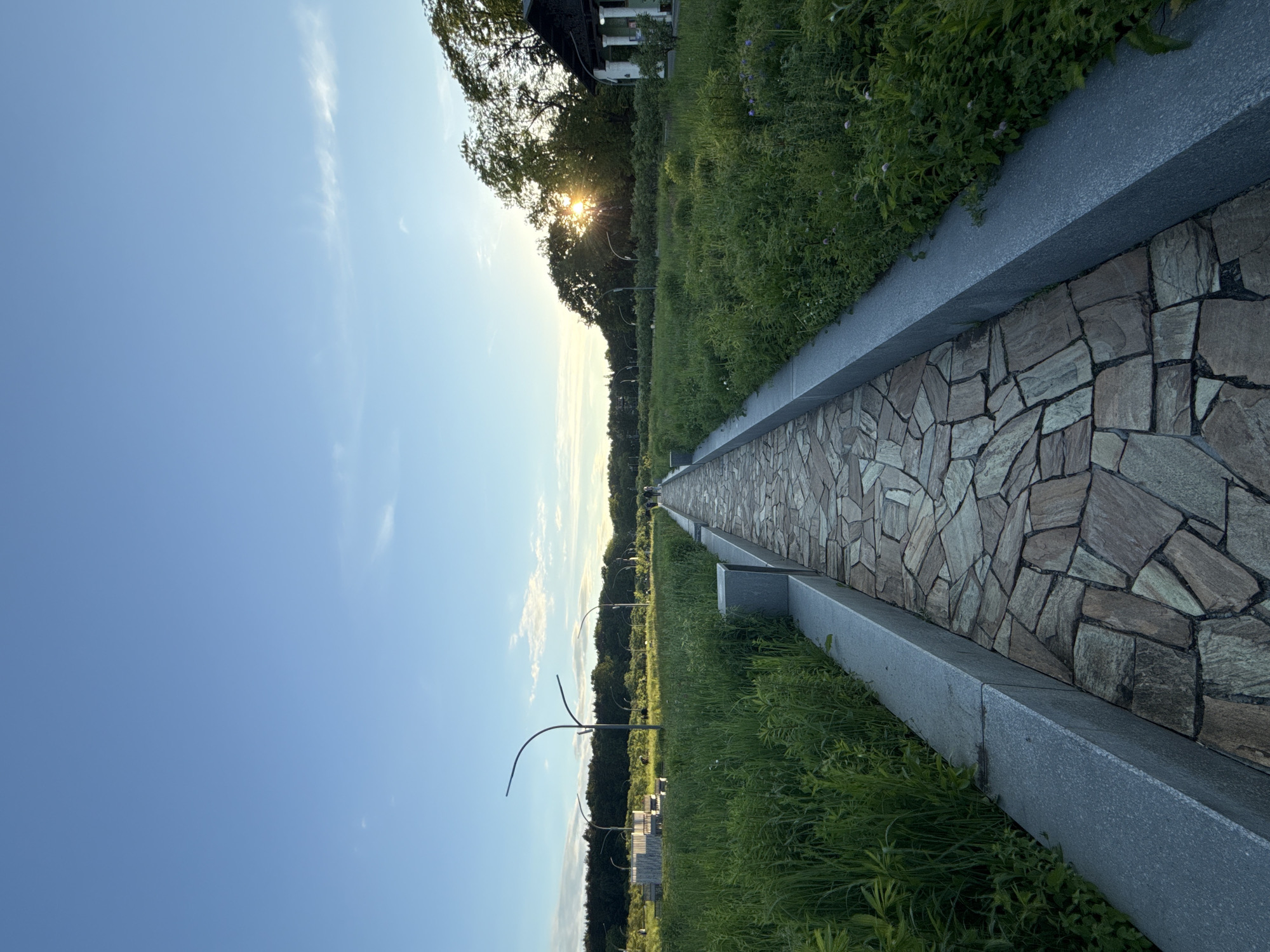 The image depicts a serene outdoor landscape, likely a park or promenade, under the warm glow of a late afternoon or early evening sun. The sun is visible on the right side, peeking through dense green tree foliage, creating a bright sunburst effect.
A long pathway, paved with irregular, multi-toned stone or tile pieces, extends from the foreground into the distance, drawing the viewer's eye. This path is bordered on its left by a solid, grey concrete or stone curb, which separates it from a broad stretch of vibrant green grass. The grass appears lush and well-maintained.
Beyond the grassy expanse, a distant treeline creates a dark silhouette against the brighter horizon. Several modern, sculptural streetlights or decorative poles with curved designs are scattered within the grassy area. In the upper right corner, partially visible behind the trees, is a section of a building featuring columns and a dark roof.
The sky is a clear, bright blue, with only a few faint, wispy clouds, indicating fine weather. The scene conveys a sense of peacefulness and calm. No people are visible, and no specific activities are taking place. There is no visible text in the image. The setting is identified as being in Moscow, Russia.
