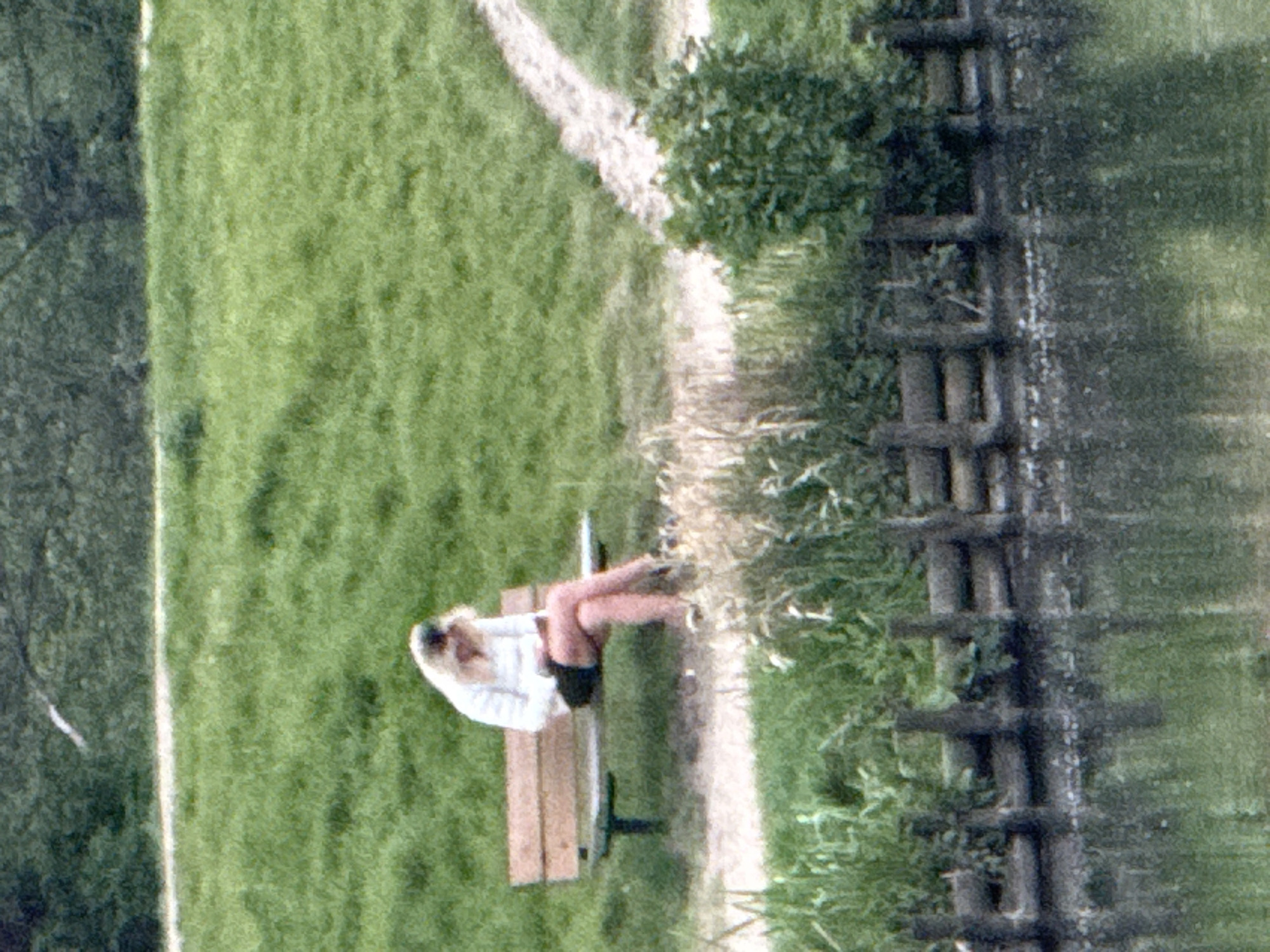 A woman sits on a park bench near a pathway and wooden fence. The location cannot be determined as London from the details shown.