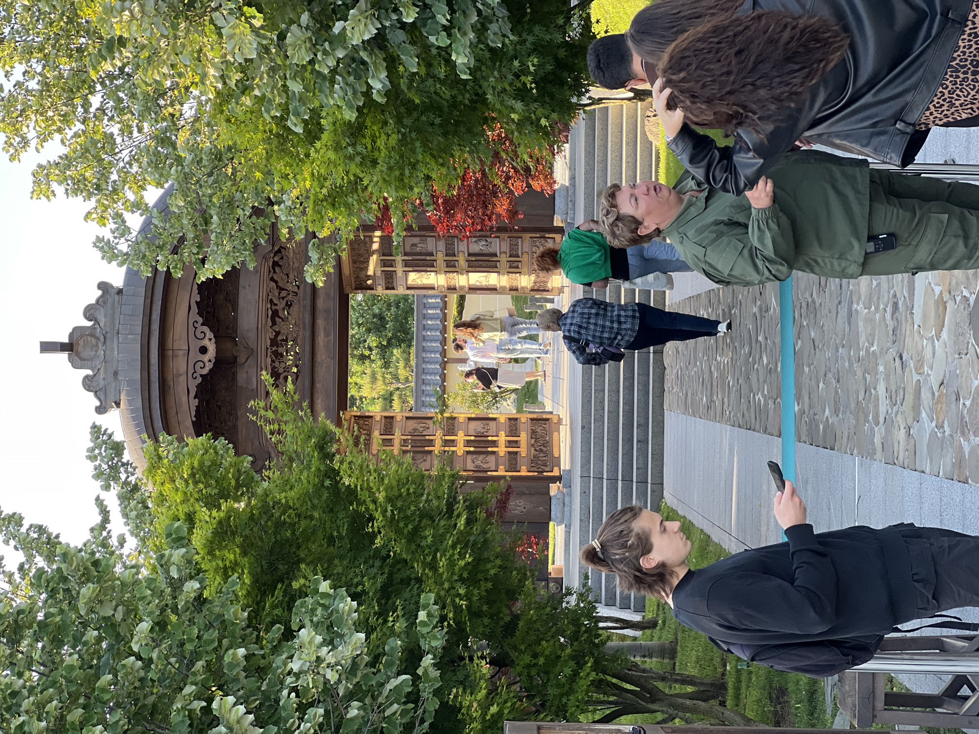 People are walking or standing in front of a traditional East Asian structure, likely a gate or pavilion. The structure is made of dark wood and adorned with intricate carvings. The architecture suggests a location with cultural ties to East Asia. Green foliage frames the structure on both sides, indicating a park or garden setting. In the foreground, a person is looking at a smartphone. Other people are interacting in small groups, some looking toward the structure. Based on the information provided, this image is set in the Japanese Garden in the city of Krasnodar, Russia.