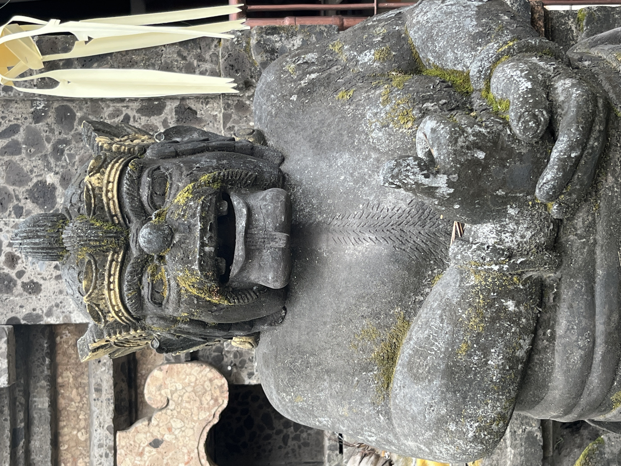 An aged, weathered stone statue of a mythical guardian figure stands prominently in front of weathered stone architecture. The statue is dark gray and covered in patches of green moss, indicating age and exposure to the elements. It features a stern, wide-mouthed expression with its mouth agape and prominent teeth. The figure wears an ornate, golden crown with a spiky crest. Its hands are clasped together, holding what appears to be a smaller object or symbol. The background consists of rough stone walls and decorative carved elements, suggesting a temple or sacred site. To the right, yellow palm leaf decorations hang vertically, and a reddish-brown metal grate is visible. The overall impression is one of ancient artistry and cultural significance, likely located in a temple complex in Kuta Selatan, Indonesia.