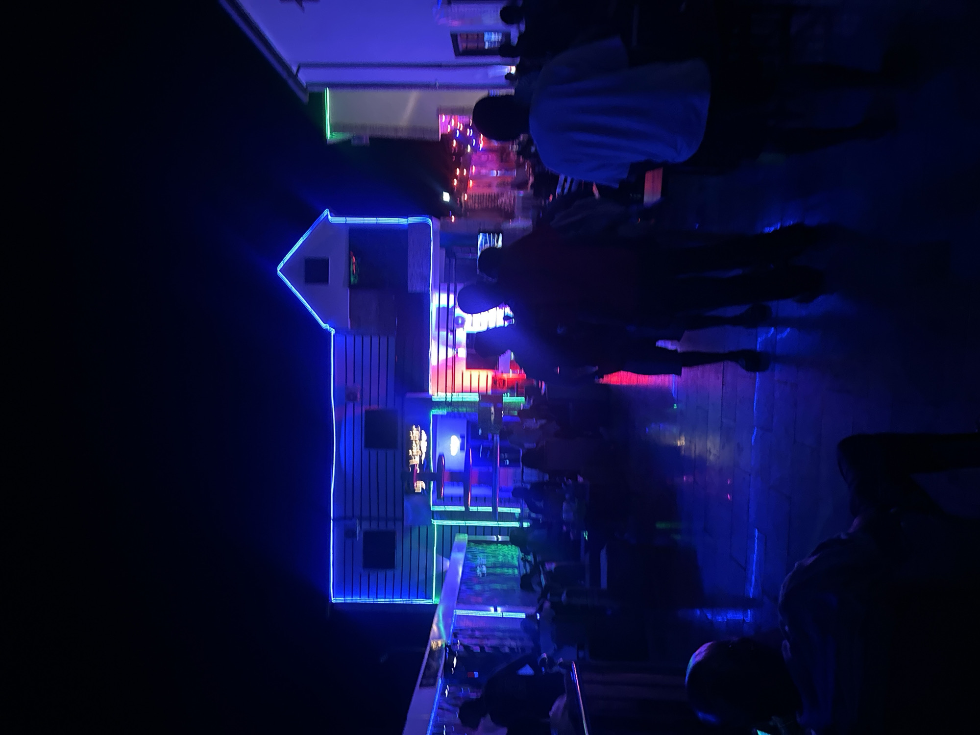 The image depicts a dimly lit outdoor scene in Tumfure, Nigeria, dominated by a two-story building outlined with bright blue neon lights. The building appears to be a commercial establishment, possibly a bar or restaurant, as indicated by the tables and chairs visible along the bottom edge of the structure. Several people are present, though their features are mostly obscured by shadow and darkness. Some individuals are standing or walking in front of the building, their silhouettes illuminated by the multi-colored neon lights emanating from within. The overall atmosphere is one of nightlife activity, with the lighting suggesting a recreational or entertainment venue. The environment is characterized by a street or pedestrian area, with other buildings visible in the background, also featuring neon lighting, further emphasizing the lively urban setting.