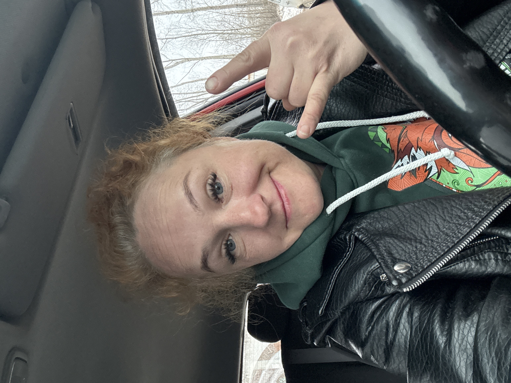 A person with red hair is sitting in a vehicle, wearing a green hoodie under a black leather jacket. They are making a hand gesture with their right hand. The environment outside appears to be wintery with bare trees. No specific details indicate the location is Chaikovsky, Russia.