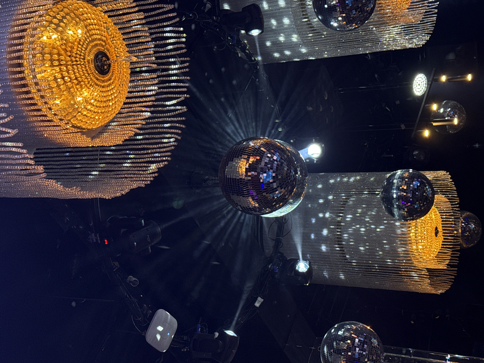 The image depicts a low-angle view of a darkened interior space, likely a nightclub or entertainment venue. The ceiling is adorned with several disco balls and elaborate chandeliers, with the light fixtures exhibiting a sparkling, crystalline appearance. At least five disco balls of varying sizes are visible, reflecting light in all directions. The chandeliers feature a complex arrangement of reflective elements, with one prominently displaying a warm, golden hue. Several spotlights and stage lighting fixtures are positioned around the ceiling, directing beams of light towards the disco balls and chandeliers. The background appears dark and undefined, suggesting a focused illumination of the decorative elements. The image captures a scene of entertainment or celebration, likely taken in Kazan, Russia.
