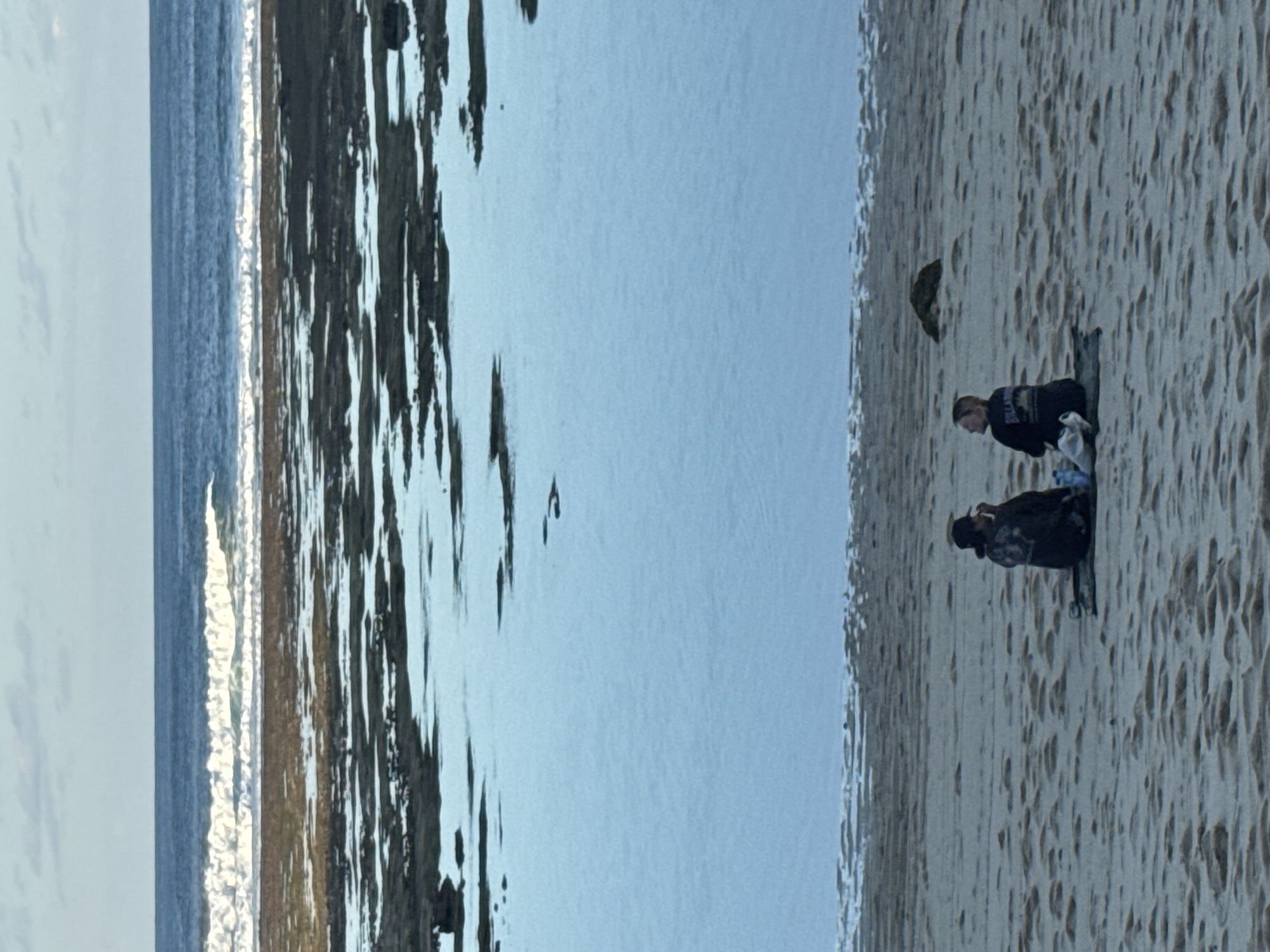 Two individuals sit on a sand blanket on a beach. The person on the left is wearing a dark jacket and a baseball cap, facing away from the camera. The person on the right is wearing a dark shirt and looking to their left. The individuals are positioned on the sand with the ocean and tide pools in the background. The ocean is a deep blue, with white waves breaking further out. The tide pools are shallow and reflect the blue of the sky and ocean. The overall scene appears to be a tranquil moment on a beach in Kuta Selatan, Indonesia.