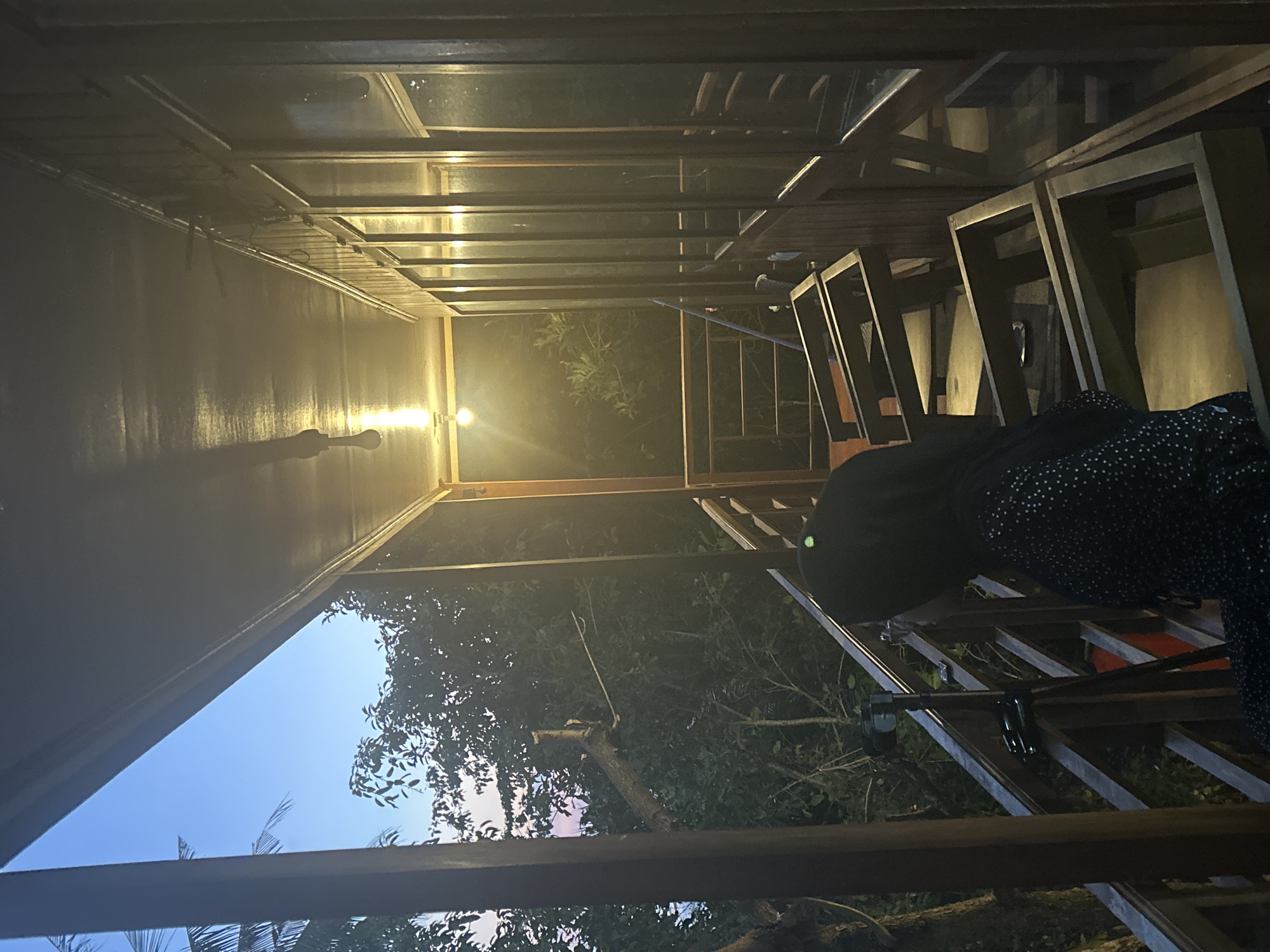 A person wearing a black dress with white polka dots is visible on the right side of the frame, appearing to be ascending a wooden staircase. The scene is outdoors, possibly a balcony or veranda of a building constructed with dark wood. The architecture features a sloped roof made of metal or glass, supported by wooden beams. Light from a bulb illuminates the left side of the scene, casting a shadow of what appears to be a broom or similar long object. Through the open structure of the building, lush green foliage and trees are visible, suggesting a tropical or semi-tropical environment. The sky in the background shows a gradient of twilight colors, transitioning from a deep blue to a lighter hue near the horizon, indicating either dawn or dusk. The overall atmosphere is serene and dimly lit, with the natural light interacting with the artificial light source. There are no visible text elements in the image. The location is specified as A R A, Indonesia.