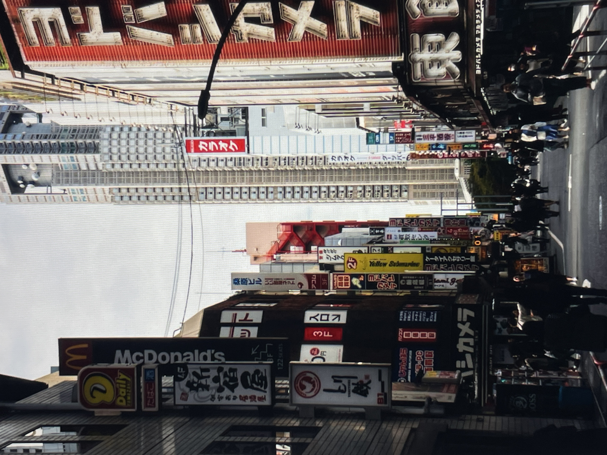 The image depicts a street scene in an unidentified Japanese city, densely populated with pedestrians on the right side of the frame. The buildings lining the street are heavily adorned with various signs, primarily in Japanese, featuring brands like McDonald's, Daily Yamazaki, Yellow Submarine, and Yodobashi Camera, indicating commercial activity. A tall, modern building is visible in the background.