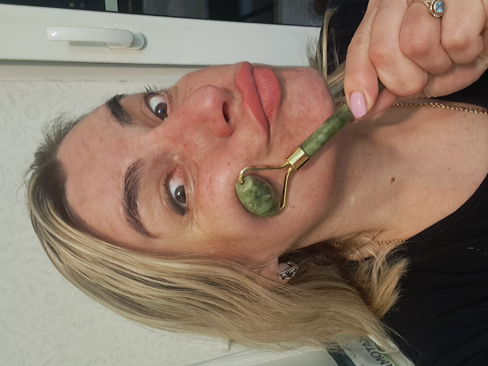 A woman is using a green jade roller on her cheek. She has blonde hair, light skin, and is wearing a ring and a gold necklace. The background is indistinct but appears to be a room with a window. There is no information to suggest this image was taken in Elektrostal, Russia.