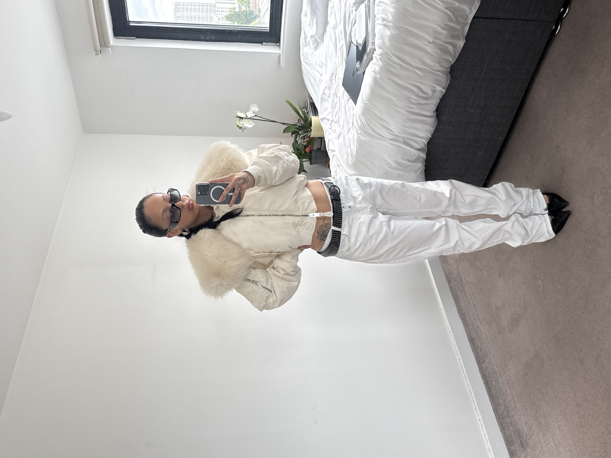 A person wearing sunglasses and dressed in white is taking a selfie in a room. The room appears to be a bedroom, with a bed covered in white bedding and a gray headboard. There is a window in the background with a view of buildings, possibly in London, United Kingdom. An orchid plant sits nearby the bed. The person is wearing a white jacket with fur trim, white pants, and a studded belt. Their pose is casual, with one hand on their hip and the other holding the phone. The walls are white, and the floor is carpeted.