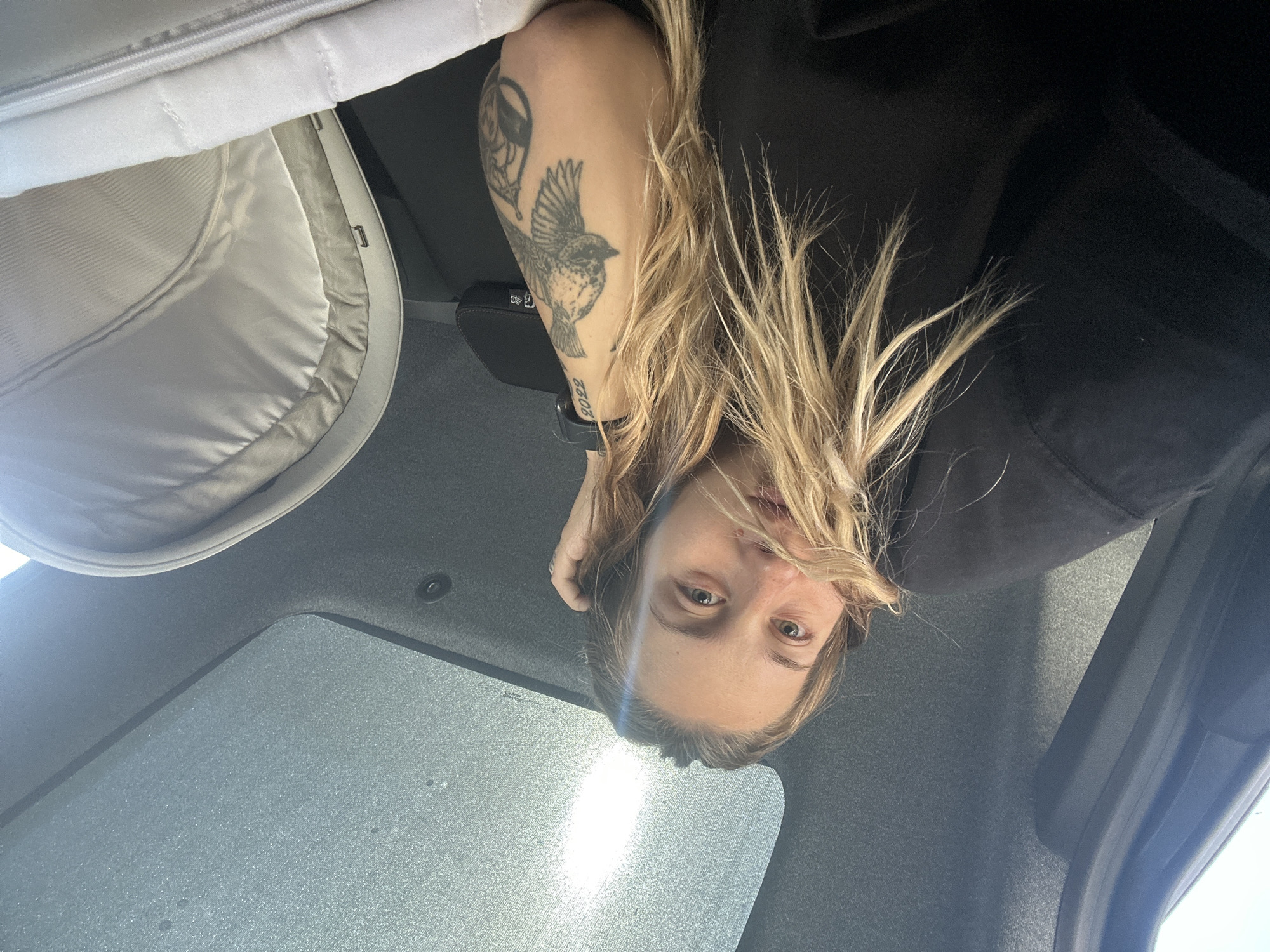 A young woman with long blonde hair and a tattoo on her arm is sitting in the back seat of a car, with her hand on her head. She is wearing a black sweatshirt. Next to her is a white baby stroller. The car has a sunroof that is open, and the sky is visible through it. The interior of the car is dark gray. The scene appears to be in a car traveling on a road, possibly in Plovdiv, Bulgaria, given the context provided.