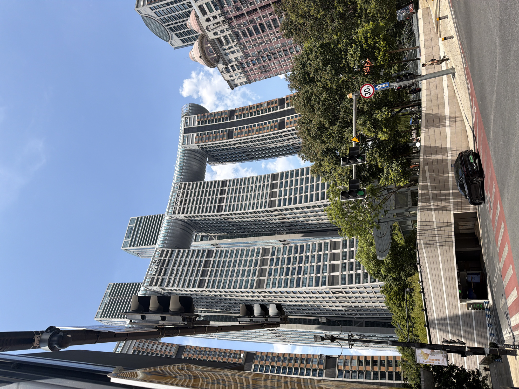 The image depicts a street-level view of modern architecture in Yuzhong District, China. Dominating the scene are several tall, contemporary buildings, including one with a unique arched structure connecting two towers at a high elevation. In the foreground, a black car is parked on the side of a road with red and white striped crosswalk markings. Traffic lights are visible, one of which shows a green signal. A street sign indicating a speed limit of 30 is also present. Several trees and lush green foliage surround the buildings and a pedestrian pathway. A person is walking along the pathway. The sky is clear and blue with some scattered white clouds. An entrance to an underground parking structure is visible in the lower portion of the image. The scene appears to be captured on a bright, sunny day.