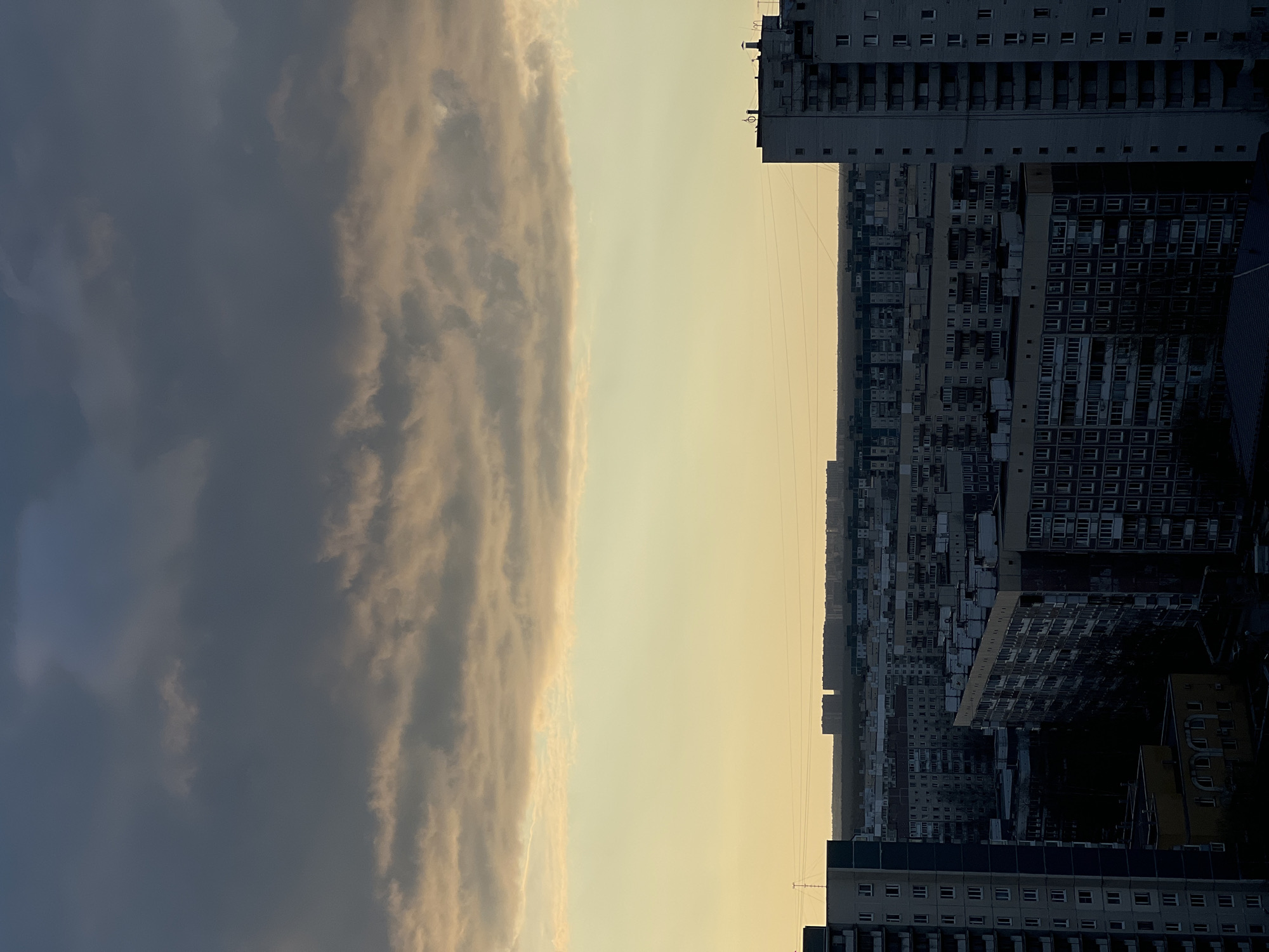 The image is an aerial shot of Saint Petersburg, Russia, at twilight, showing a cityscape with residential buildings under a cloudy sky. The buildings are predominantly gray and display a repetitive, grid-like facade with numerous windows. The sky is divided into a dark, overcast area and a brighter, golden-hued section. There are no visible people or specific interactions. Electrical wires are also present in the sky.