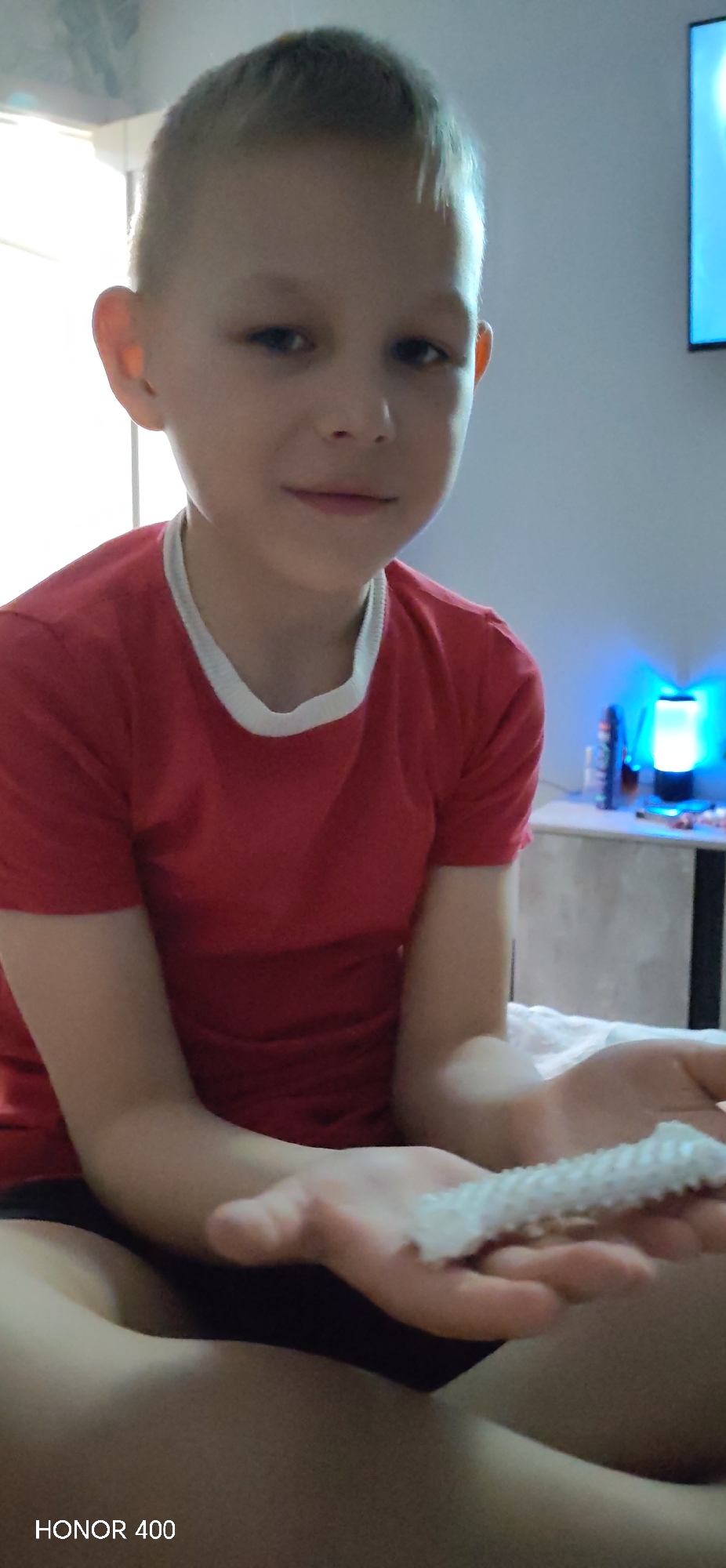 A boy with short blonde hair is seated, wearing a red T-shirt with a white collar, and holding a white textured object in his hands. Behind him is a bedside table with a blue lamp and a TV mounted on the wall. The location appears to be inside a residential room. There is no verifiable location in Noviy, Russia visible.