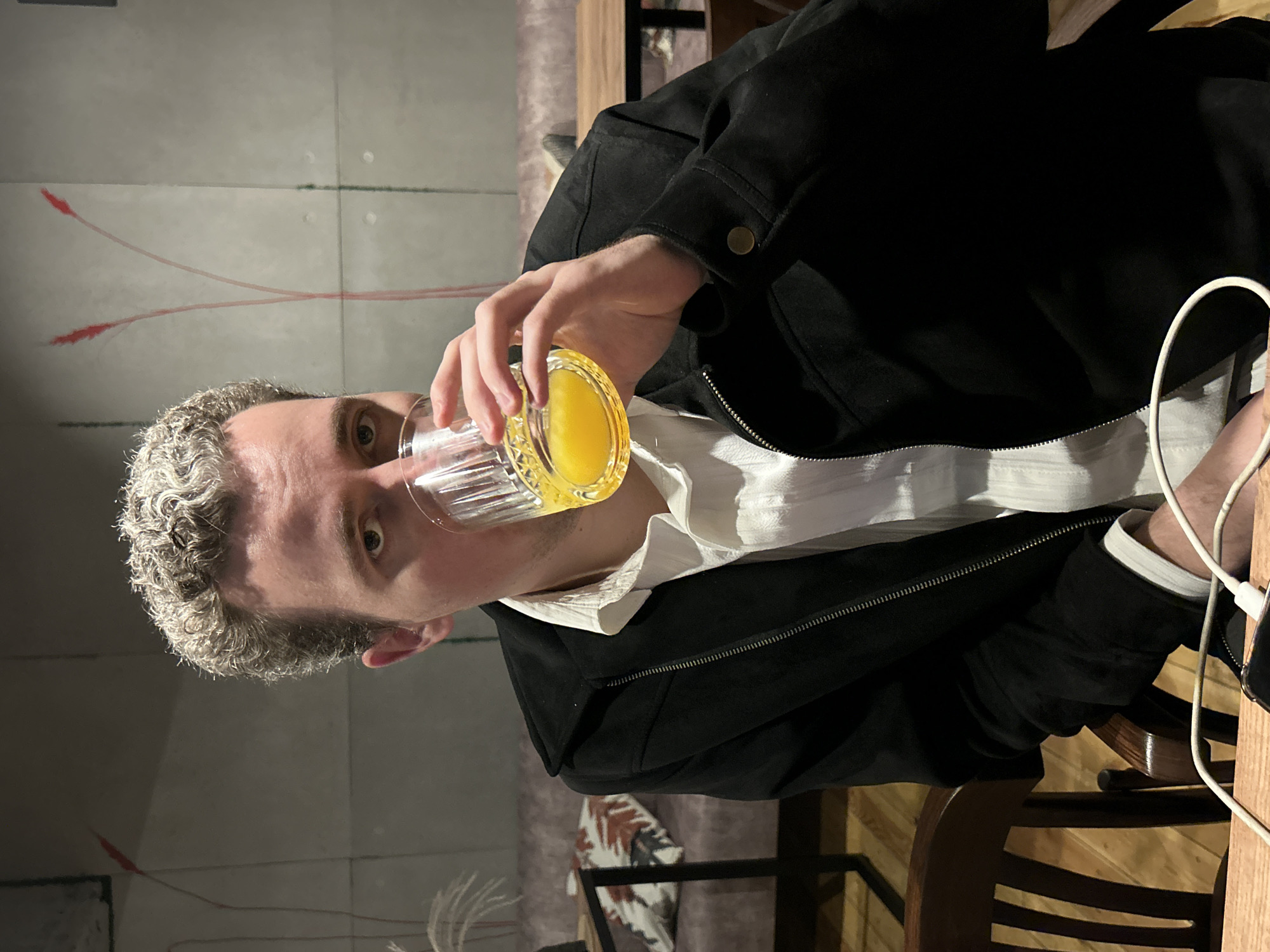 A man with light curly hair is sitting at a table and drinking from a glass of orange juice. He is wearing a white shirt and a black jacket. In the background, there is a sofa and a table. The location is not Saint Petersburg, Russia.