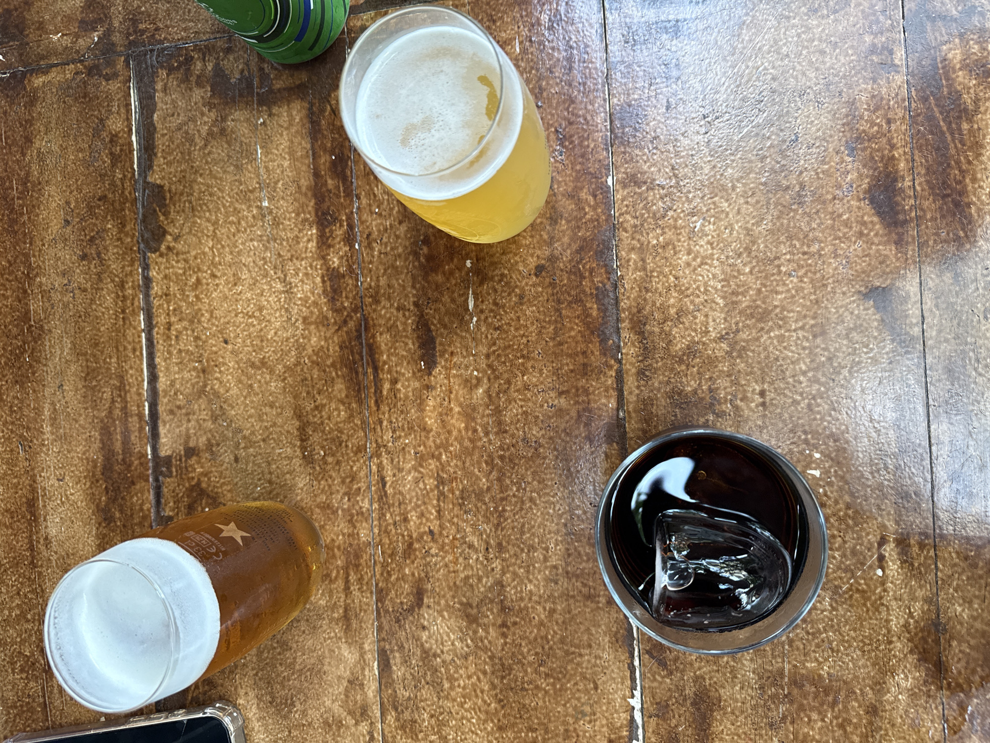 An overhead photograph captures three beverages positioned on a worn, multi-planked wooden table. In the upper-left quadrant, a clear glass contains an amber-colored beer topped with a substantial white foam head. The glass is visibly marked with "33 cl", "CE E8", and a distinct five-pointed star logo, consistent with Estrella Damm beer, a product widely associated with Barcelona, Spain. Faint text including "RITZENHOFF" is also discernible on the glass. A smartphone encased in a transparent cover is partially visible along the far-left edge of the frame. In the lower-center of the image, a second clear glass holds a dark brown liquid containing a single, large, irregularly shaped ice cube. To the mid-right, a third clear glass contains a pale yellow beverage crowned with a thin layer of white foam. A segment of a green object, possibly a bottle or can with blue and black striped markings, is visible in the upper-right corner. The wooden table surface displays prominent grain, variations in brown tones, and visible signs of wear and age, consistent with a used surface. No individuals are depicted in the scene.