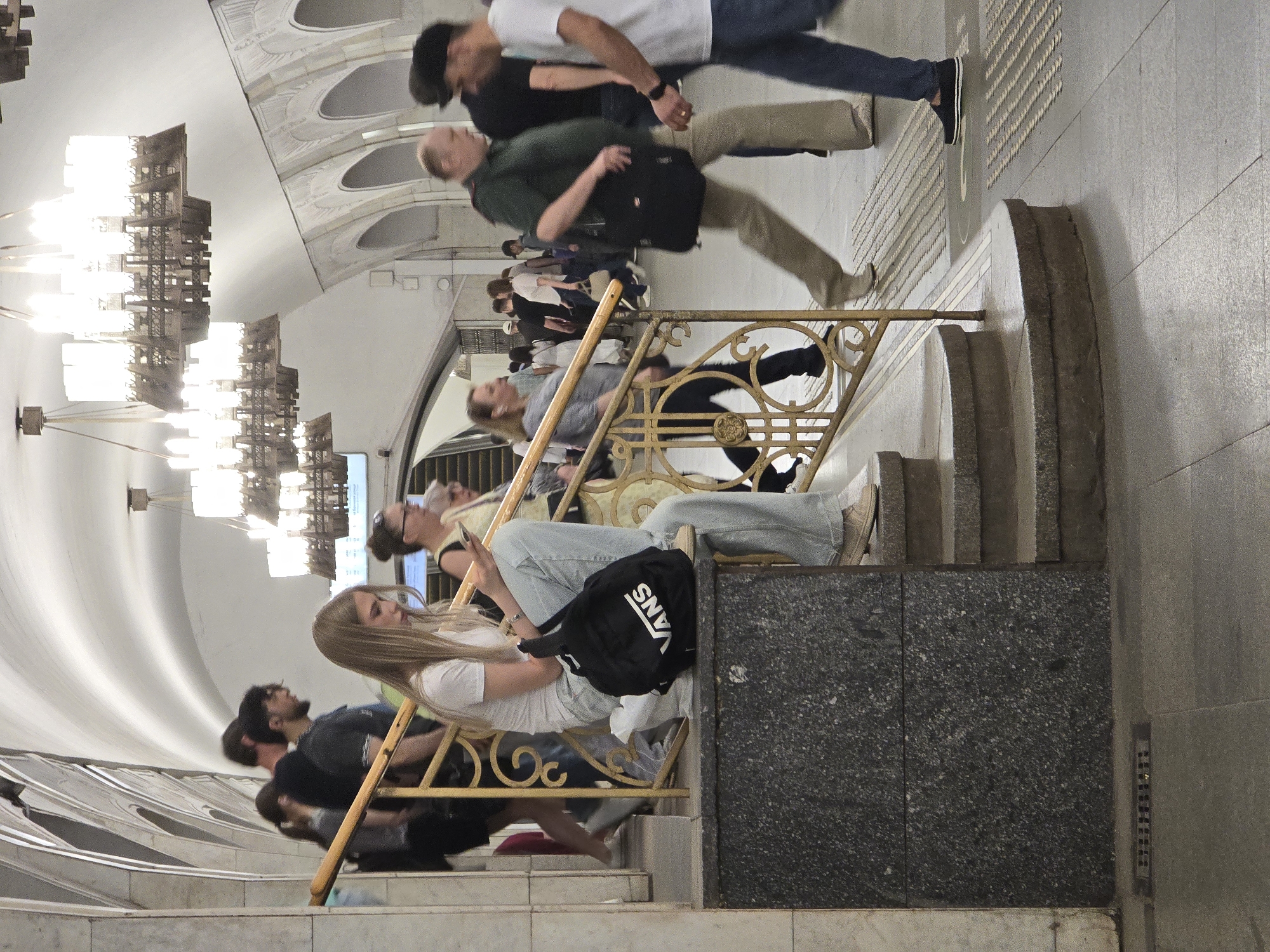 The image shows an interior scene, seemingly a subway station or public transportation hub, filled with people. The walls and ceiling are adorned with elaborate architectural details, including arched forms and decorative lighting fixtures. Several people are visible in the scene, some walking along a tiled floor, and others sitting on or near the railings of a staircase. The attire of the individuals is varied, suggesting a mix of ages and styles. A large black backpack with the word "VANS" is prominently displayed. The setting suggests a station or similar public space, perhaps in an urban area.
