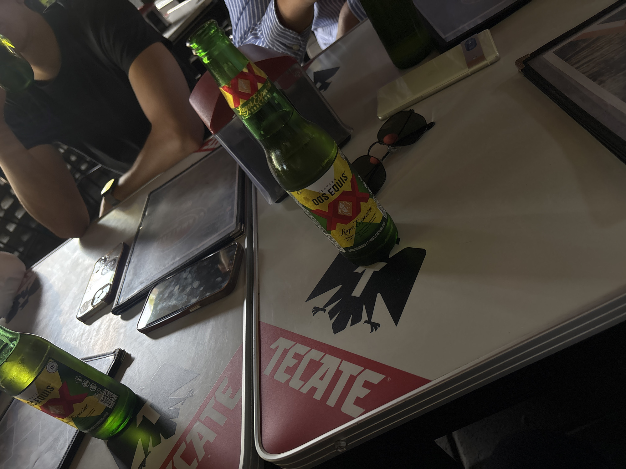 The image depicts a table with a Tecate logo. On the table are several items, including Dos Equis beer bottles, phones, sunglasses, and a wallet. People are partially visible around the table. The location is not identifiable in the image.