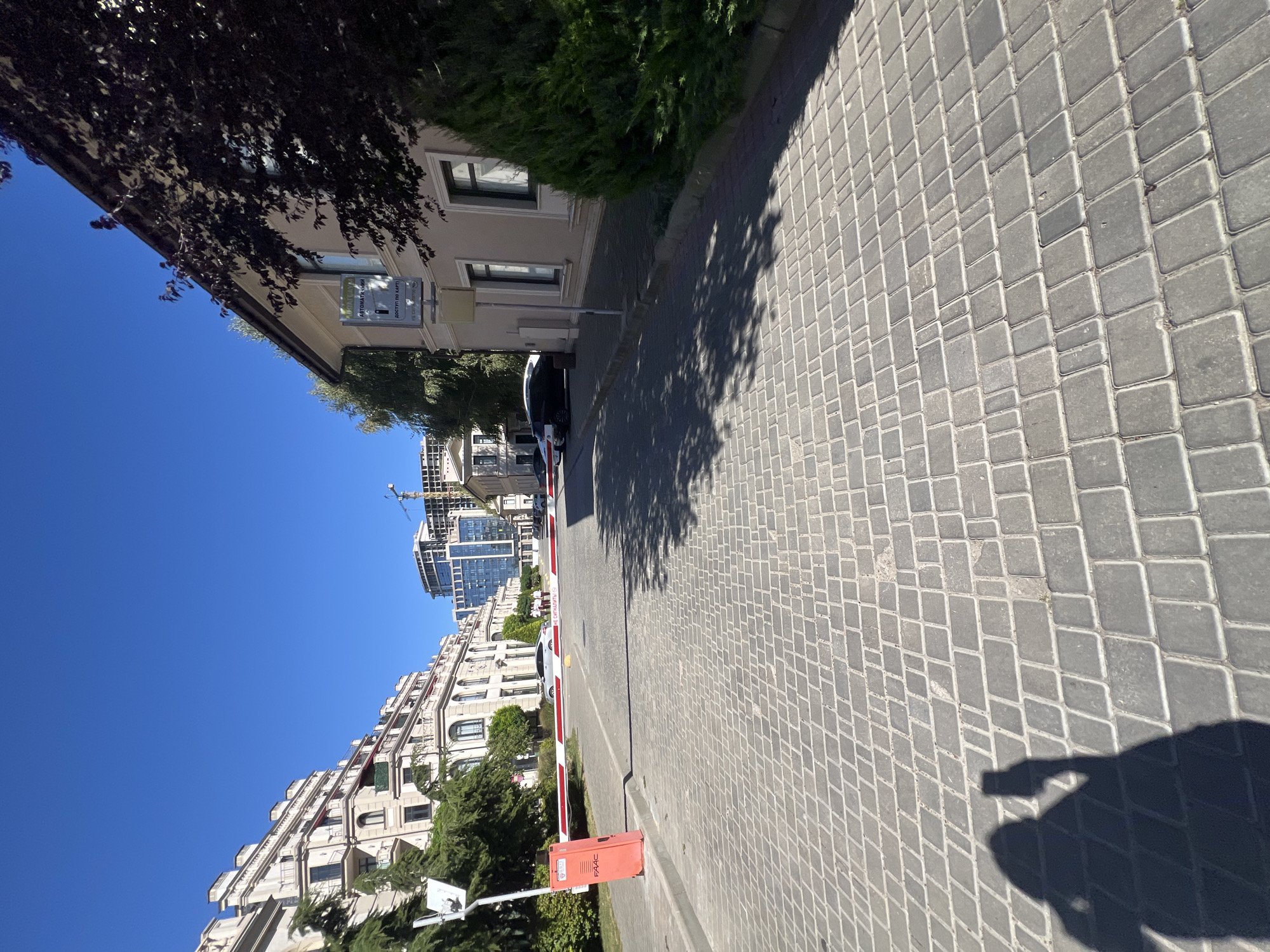 This is a sunny, exterior, eye-level, medium shot of a paved road with a red and white barrier. The barrier is located at an entrance, with a red FAAC-branded machine on the left side. Beyond the barrier, the road extends towards a row of buildings. On the left, there's a large, ornate cream-colored building with multiple balconies and arched windows, partially obscured by green trees. In the distance, modern buildings, some under construction with cranes visible, rise against a clear blue sky. On the right side of the road, a lighter-colored building with dark-trimmed windows stands. A mature dark-leaved tree partially shades the right side of the scene. The ground is paved with grey rectangular stones laid in a repeating pattern. In the foreground, a dark silhouette of a person holding a phone is cast on the pavement, suggesting they are taking a photo or recording a video. The scene appears to be in a developed area, possibly a residential or commercial complex in Fontanka, Ukraine.