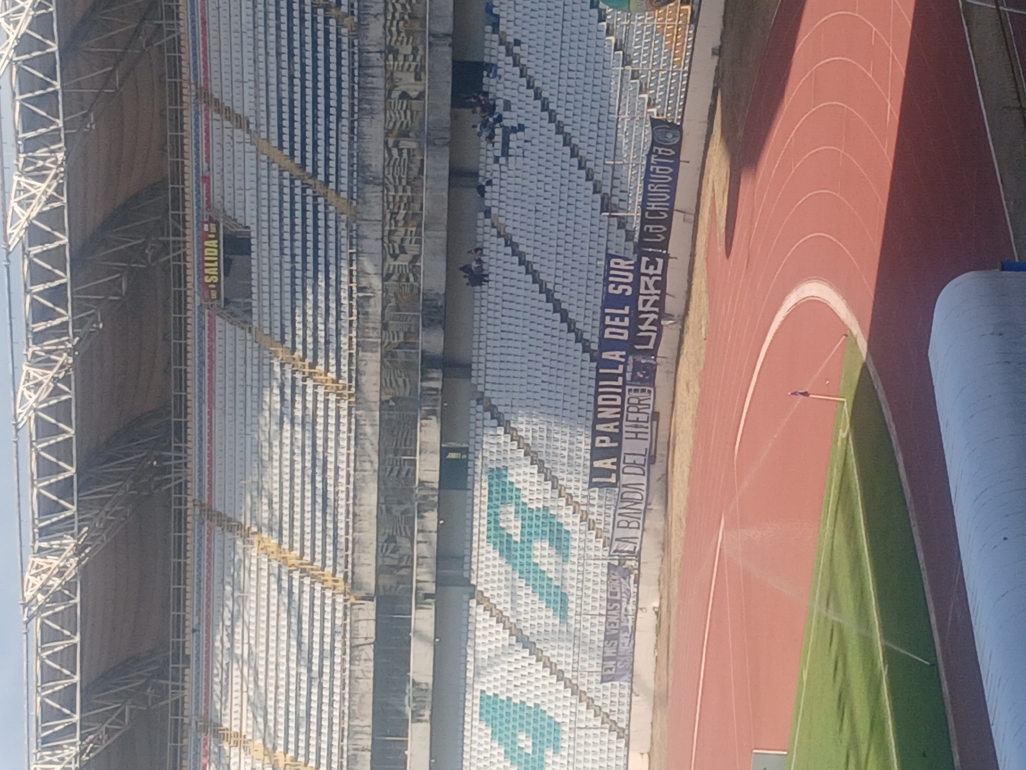 The image shows a view inside a stadium. In the background, stadium seating stretches across the frame with alternating sections of empty white seats and blue and white patterned seats, along with some with writing and symbols. A red running track is visible on the right, adjacent to a green field. In the top left of the image, a metallic framework supports the stadium's roof. A banner with text is displayed near the running track, on the lower right end of the seats. The stadium is likely located in Ciudad Guayana, Venezuela.