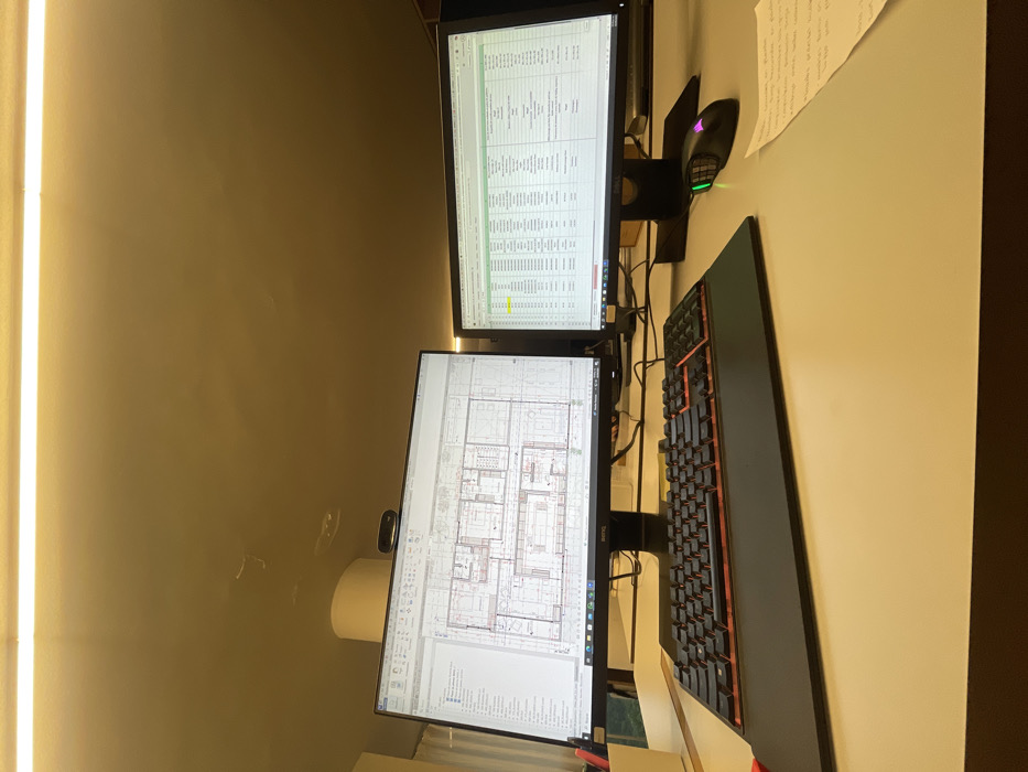 The image displays an unoccupied office workstation located in Mexico City, Mexico. A light-colored desk supports two black computer monitors. The left monitor, a BenQ, exhibits architectural floor plans and user interface elements from design software. The right monitor, a Dell, shows a spreadsheet with multiple columns of data, including yellow-highlighted cells. Directly in front of the monitors, a black mechanical keyboard with orange-backlit keys rests on a wrist support. To its right, a black gaming mouse with green and pink/purple LED lighting is positioned. A small, crumpled piece of paper with handwritten notes in Spanish is visible on the desk to the right of the mouse. A webcam is mounted atop the left monitor. In the background, a white cylindrical structure is partially visible behind the left monitor. The scene is illuminated by a prominent overhead linear LED light fixture, casting a warm yellow light across the room. No individuals are present in the image.