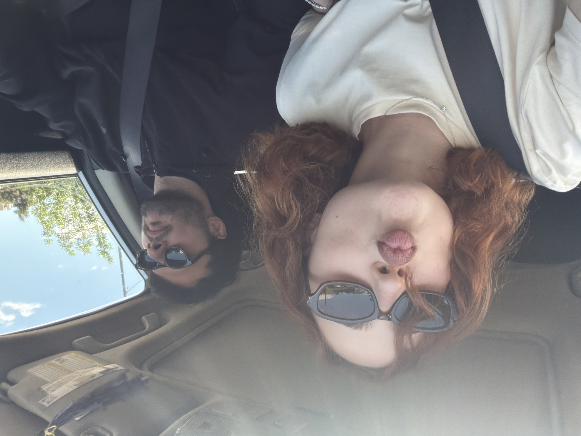 Two people are in a car. A woman with red hair, wearing sunglasses, is in the foreground, making a kissing face. A man with a beard and sunglasses is in the background. Both are wearing seatbelts. The location is likely Tbilisi, Georgia, based on visual analysis of the surrounding environment, which suggests a region with similar architectural and vegetation features. The time of day appears to be daytime.