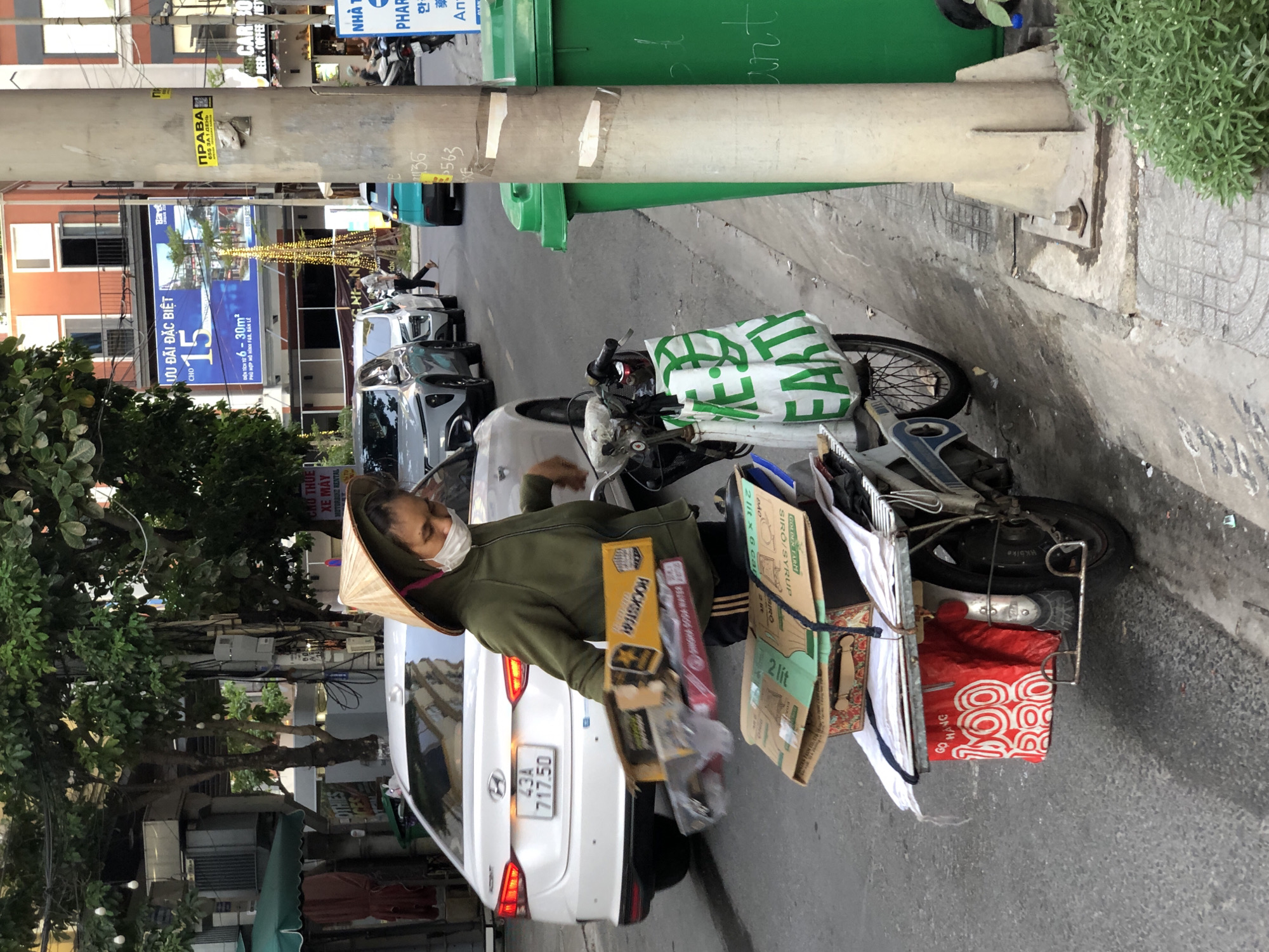 A woman wearing a conical hat and a face mask is pushing a motorcycle loaded with packages. The motorcycle is parked on the side of a street in Da Nang, Vietnam. The woman is wearing a green hoodie and black pants. She is holding a white package in her right hand and the handlebars of the motorcycle in her left hand. The motorcycle is loaded with several cardboard boxes, a red bag, and a white bag with green lettering. In the background, there are cars, buildings, and trees. The street is paved and there is a sidewalk on the right side of the street. There are also two large green trash cans on the sidewalk. The buildings in the background appear to be shops and apartments. There are also some streetlights and traffic lights visible. The overall scene depicts a busy street in a Vietnamese city.