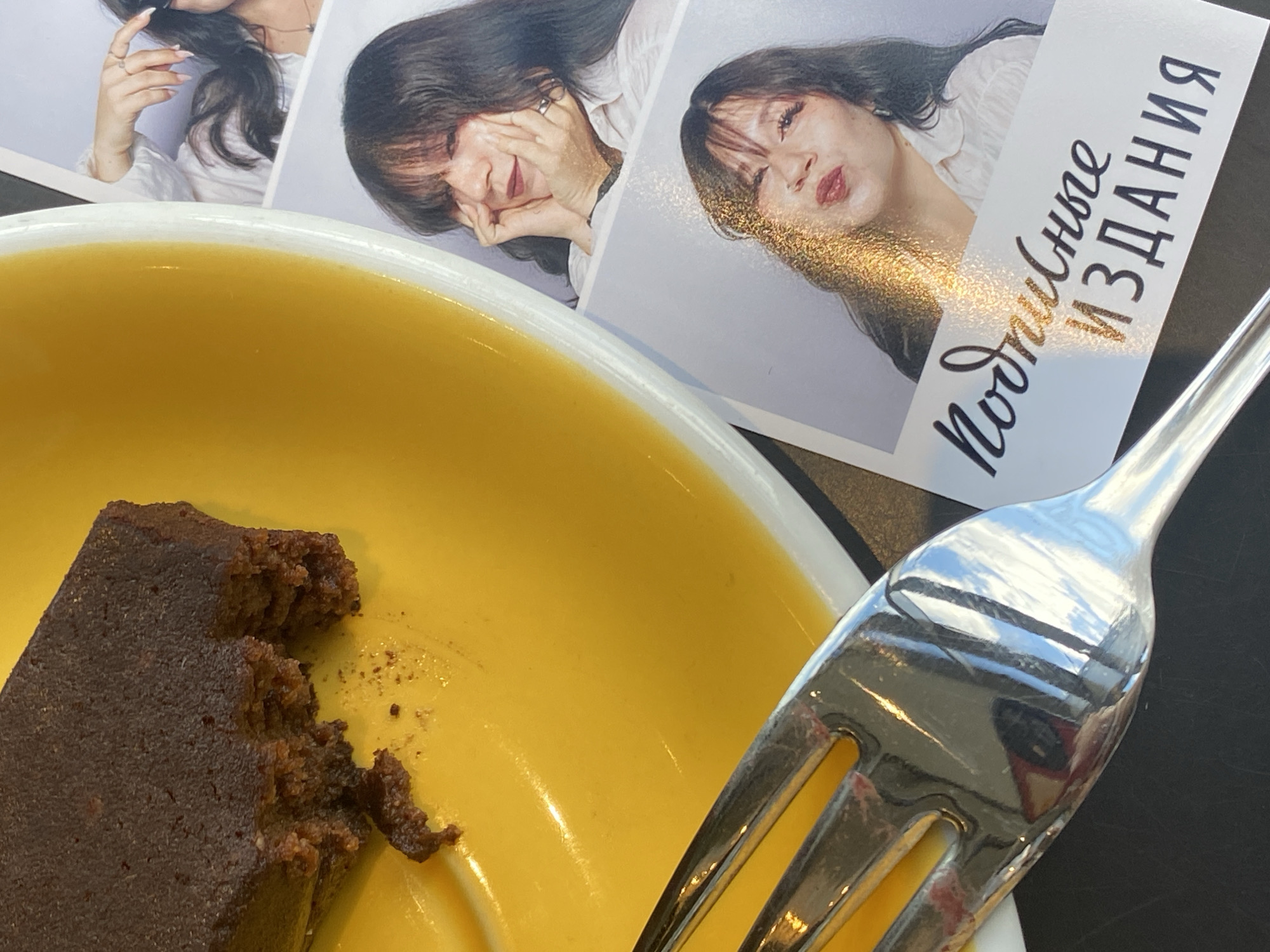 The image shows a close-up view of a piece of dark chocolate cake on a yellow plate, with a fork in the foreground. To the right of the plate are two photo booth prints of a woman. The top print shows the woman with her hands on her cheeks. The bottom print shows the woman pouting with her hair falling over her face. Text in Cyrillic script appears on a banner across the bottom of the photo prints, which reads "Подписные ИЗДАНИЯ," translating to "Subscription Editions." The setting appears to be a cafe or restaurant.