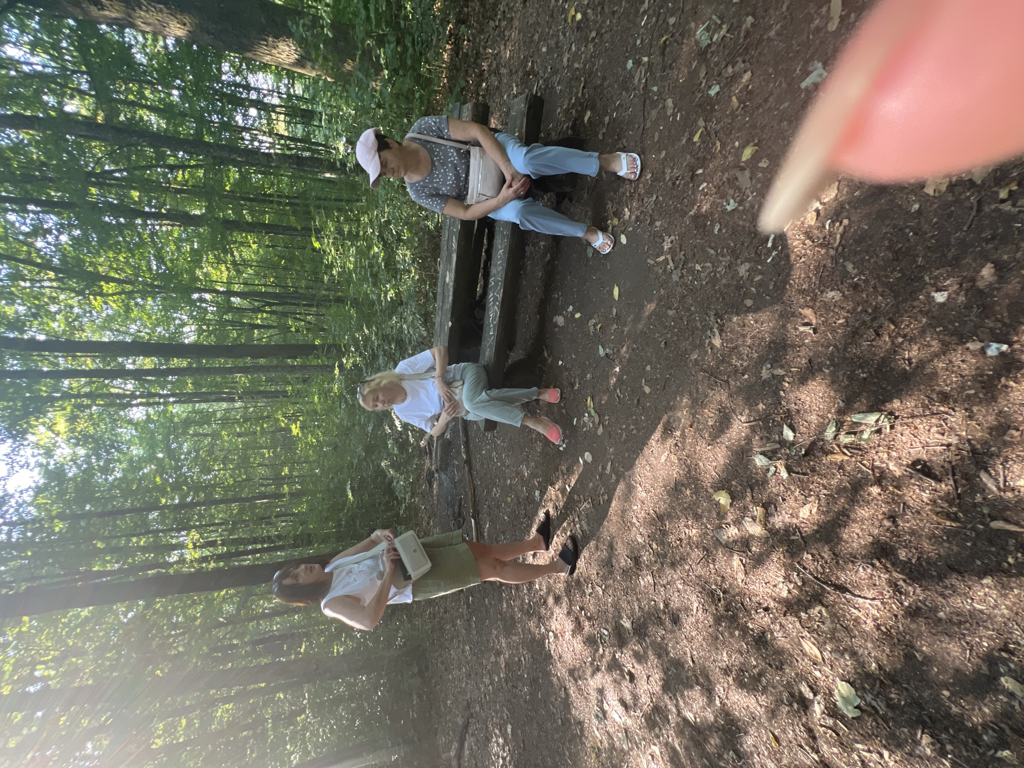 Three women are gathered in a wooded area with dappled sunlight. One woman stands on the left, wearing a white sleeveless top and olive green shorts, holding a light-colored crossbody bag. Another woman sits in the center on a wooden bench, wearing a white t-shirt and light-colored pants, with a similar crossbody bag. The third woman is on the right, seated on the same bench, wearing a grey patterned t-shirt, light blue pants, and a pink baseball cap. She also has a light-colored crossbody bag. The bench has some faint markings on it. The ground is covered with dirt, leaves, and twigs. The background is filled with tall trees and green foliage. The scene suggests a casual outdoor gathering or a break during a walk in the woods. The location is identified as Сонячне, Ukraine.