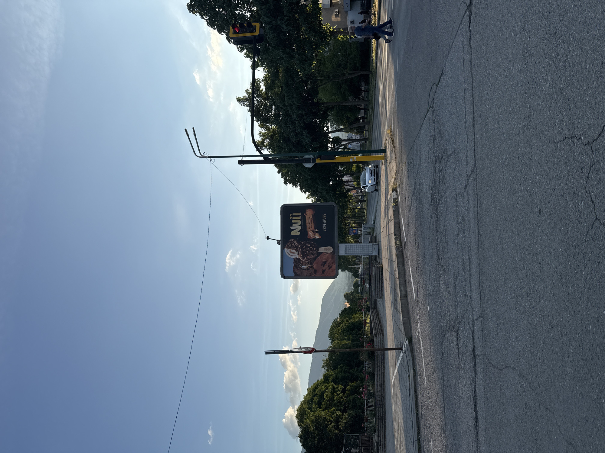 This image depicts an urban intersection in the city of Kyustendil, Bulgaria, under a clear blue sky with scattered white clouds. The sun, positioned to the right, casts long shadows.
In the center-right of the frame, a large billboard advertises "Nuii" ice cream, with "Australian Macadamia" text visible. To its right, a tall streetlight post with a green pole supports a traffic light showing a red signal for the intersecting street. Further right, two male pedestrians are captured mid-stride, walking across a paved road, from the right side of the frame towards the left, with their backs mostly to the camera. They are dressed casually in jeans and t-shirts.
On the left side of the intersection, a dense line of green trees forms a boundary for a park or green space, with stone planters and low walls visible along its edge. Distant mountains are discernible on the horizon beneath the sky in the upper-left quadrant. A single white car, possibly a sedan, is observed driving on the main road in the mid-ground, traveling from left to right. Overhead power lines traverse the sky from left to right. The asphalt road in the foreground exhibits numerous cracks, and a white line marking a crosswalk is partially visible on the right. A dark utility pole with a yellow lower section stands near the traffic light, and an additional, simpler streetlight fixture is visible to the left.