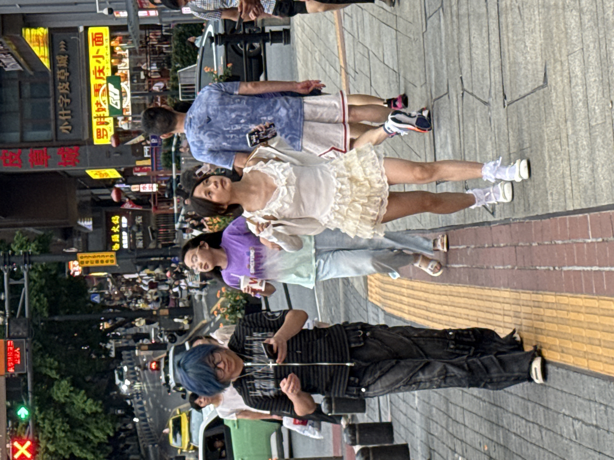 In Yuzhong District, China, a street scene unfolds with several individuals walking on a pedestrian walkway. In the foreground, a person with bright blue hair and glasses, wearing a black and grey striped zippered hoodie and dark, baggy pants, is looking down at their mobile phone. To their right, a young woman in a light purple and green ombre t-shirt and light blue jeans is holding a drink. Further to the right, another young woman is walking in a white, ruffled dress with sheer sleeves and white socks with white shoes. Behind them, a man in a blue graphic t-shirt and white shorts, and another person in a plaid shirt and shorts are also visible. The environment includes a street with traffic, buildings with illuminated signs, and trees. The walkway has textured paving.