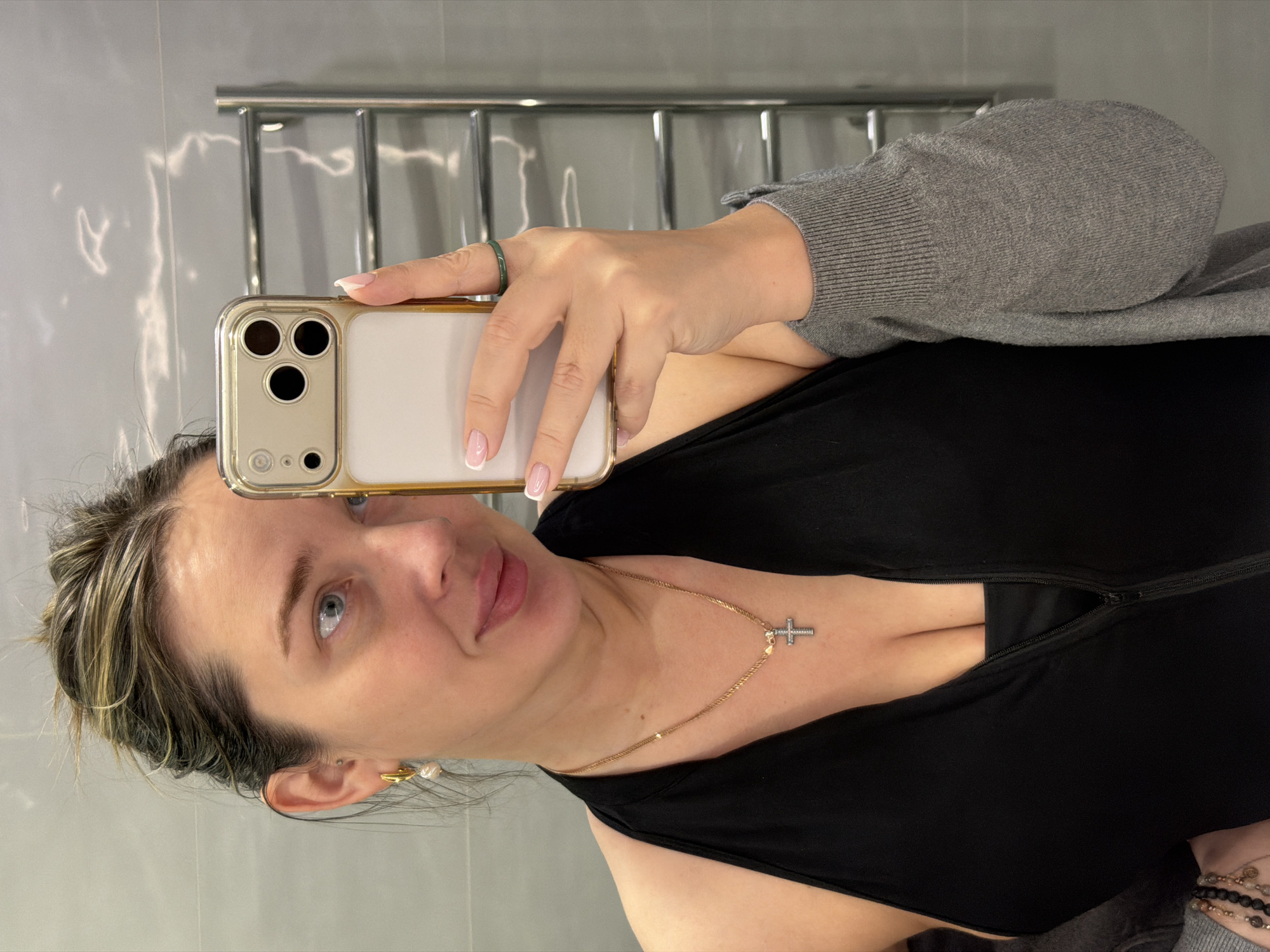 A woman is taking a selfie indoors. She is holding a gold iPhone, and wearing a black top with a low neckline, a grey cardigan, a gold necklace with a cross pendant, earrings, and a ring on her finger. The background shows a grey tiled wall and a chrome towel rack. The specific location in Dolgoprudny, Russia, cannot be determined from the image.
