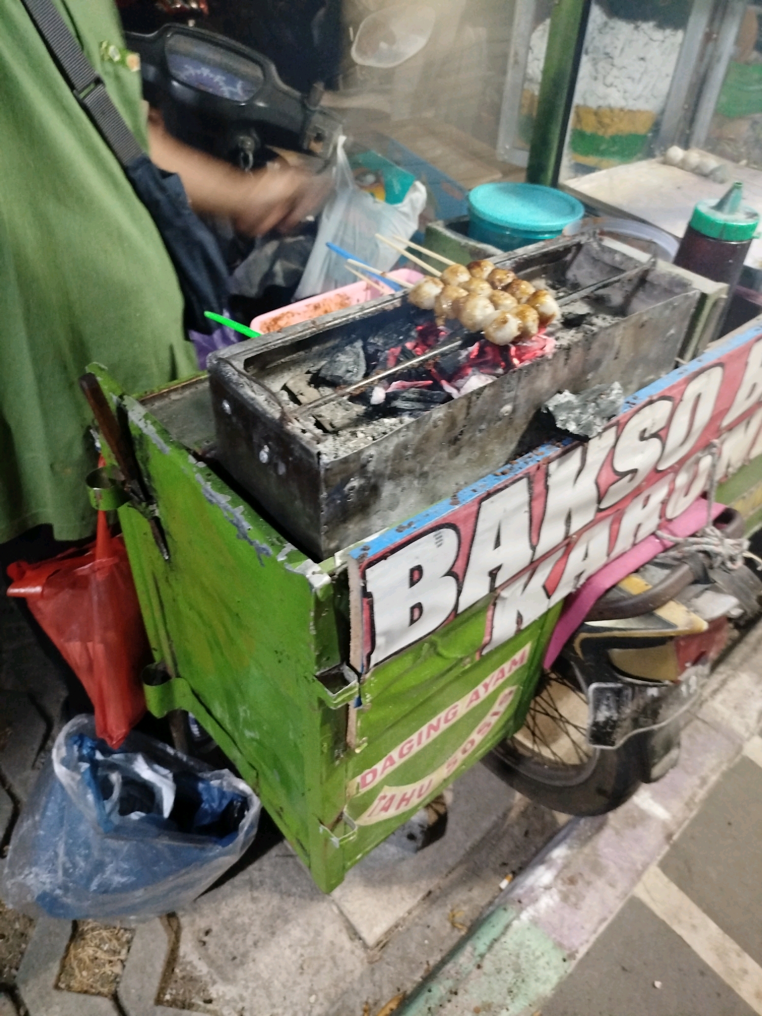 A street food vendor is preparing bakso bakar (grilled meatballs) at a mobile cart in Wonosari, Indonesia. The cart is green and features a prominent sign that reads "BAKSO BAKAR." A person, wearing a green shirt, is partially visible, tending to the grill. Skewers of meatballs are cooking over hot coals. Various containers and plastic bags are present around the cart, including a blue plastic bag on the ground and red plastic bags hanging from the cart. The scene is set on a paved street, suggesting a public urban area.