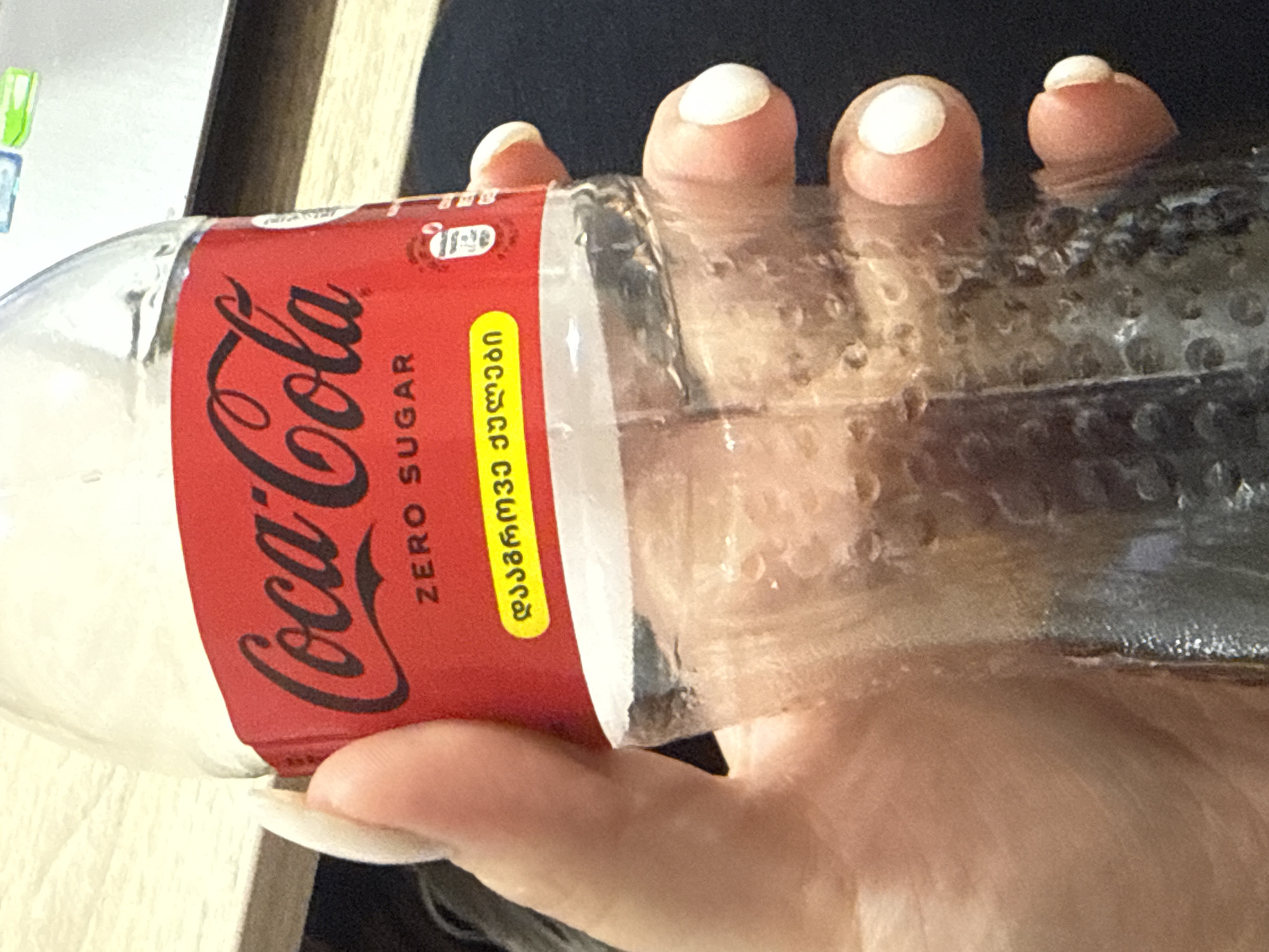 A hand with long fingernails holds a clear glass bottle of Coca-Cola Zero Sugar. The bottle has a red label with the Coca-Cola logo in black script. Below the logo, it says "ZERO SUGAR" in white text. There is also text in a yellow oval in Georgian script. The bottle appears to contain a dark liquid, possibly Coca-Cola, with some condensation on the outside. The background is blurred but suggests an indoor environment with a wooden surface and part of a laptop. The scene is captured from a slightly low angle.