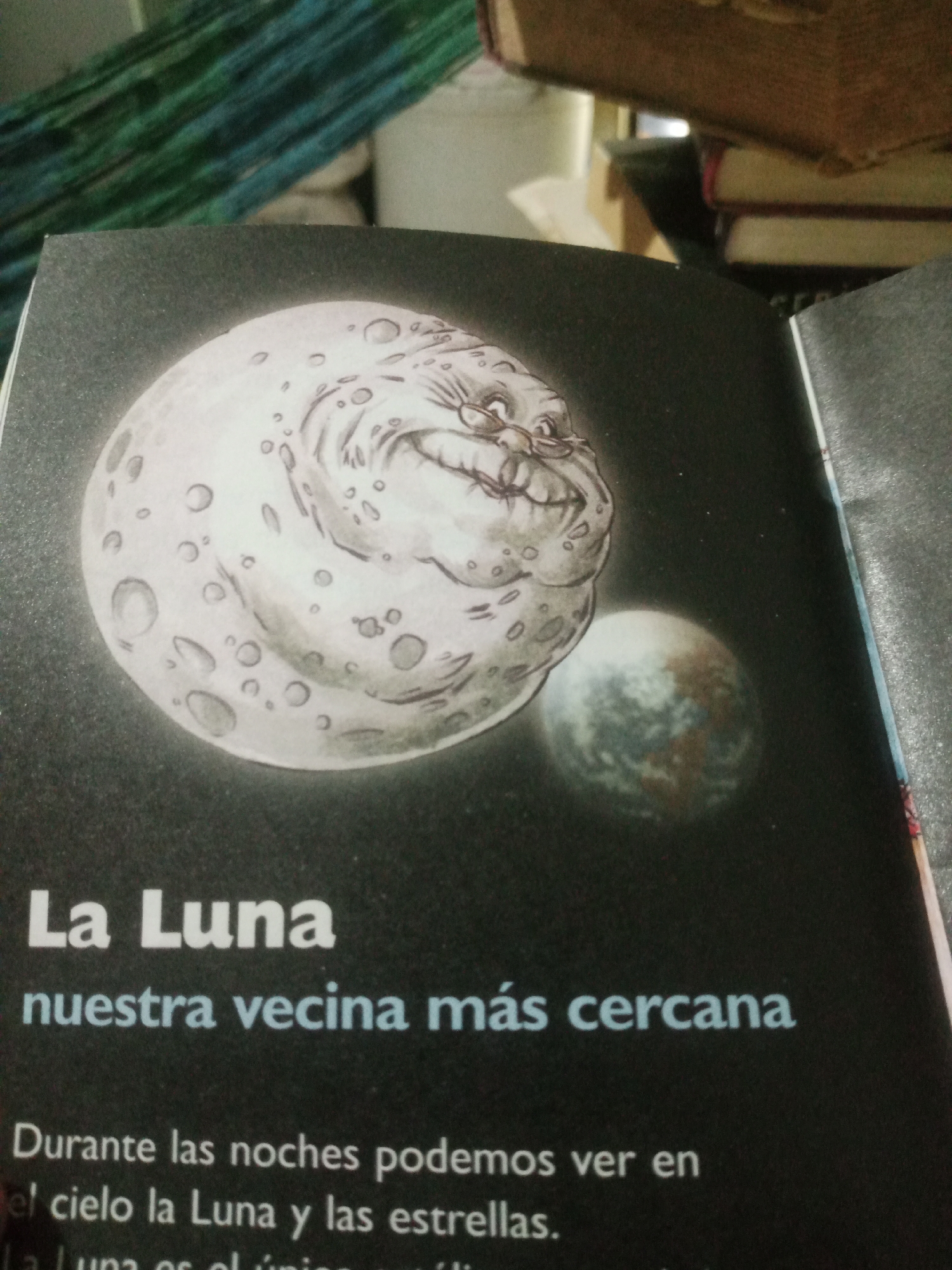 This image is a close-up shot of an open book, likely a children's book, with a dark background simulating space. The book features an illustration of a cartoonish, grumpy-looking moon with a face. To the right of the large moon is a smaller, more realistic depiction of Earth. The text on the page, in Spanish, reads "La Luna" (The Moon) and "nuestra vecina más cercana" (our closest neighbor). Below this, more text describes seeing the moon and stars at night. The book is placed on a textured, dark gray surface, possibly a table. In the background, out of focus, are stacked books with brown covers and a dark green and blue striped fabric. The image does not provide any information or context about the city of Santa Ana, Venezuela.