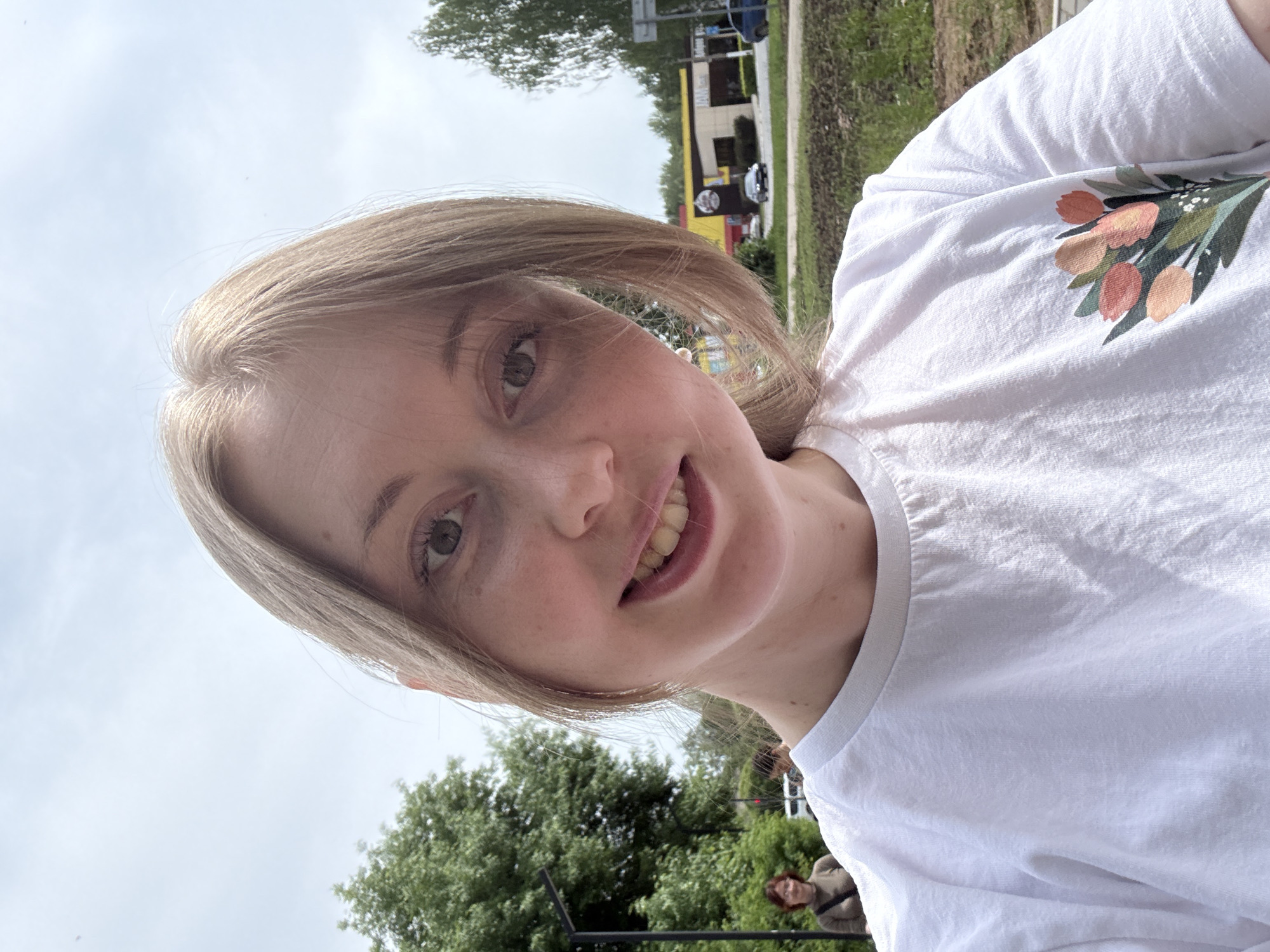 A young, light-skinned individual with light blonde hair is the primary subject of this selfie. The person is smiling and wearing a white t-shirt with a floral design on the shoulder. The background shows a tree and a building painted with yellow, red, and white stripes, possibly a commercial establishment. To the right of the background there is an unidentifiable structure. There are other people in the image, who are smaller and further in the background. The setting appears to be outdoors, possibly a park or a city street.