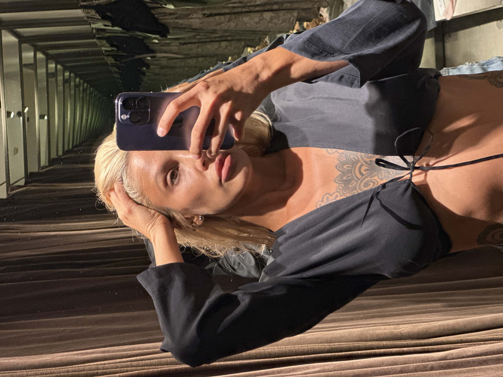 A blonde woman stands in a changing room, holding a phone to take a selfie. She is wearing a dark, cropped blouse with a tie in the front and denim shorts. Her hair is pulled back, and she has visible tattoos on her chest and arm. The changing room has brown curtains and a long mirror reflecting more changing rooms and clothing racks. The lighting suggests it is indoors.