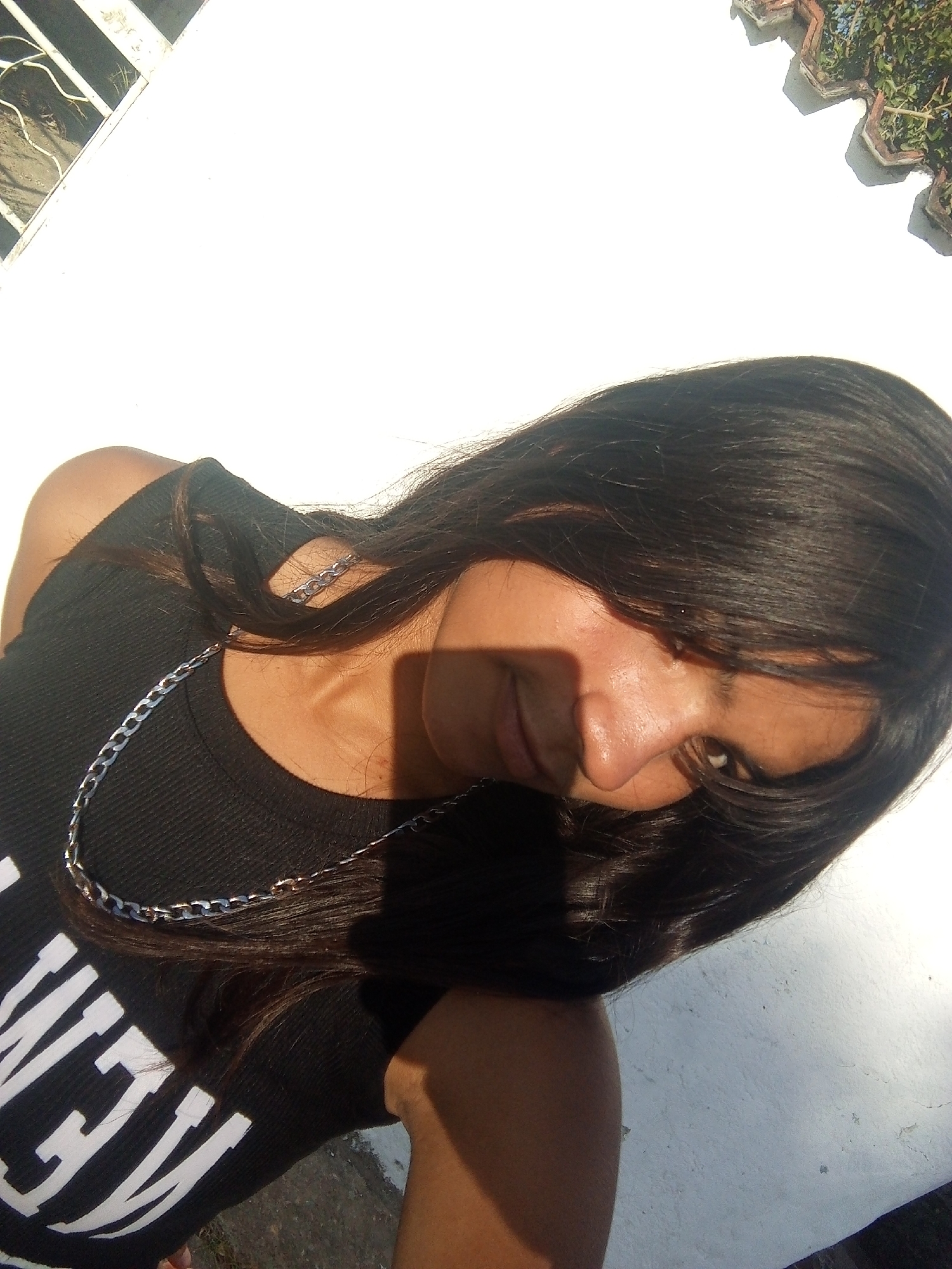 A tilted close-up shot features a young woman with long, dark hair. She is wearing a black tank top with white lettering and a silver chain necklace. The background appears to be a white wall, possibly outdoors, given the presence of shadows and what seems to be foliage visible near the top edge. The location is somewhere in the city of Juan Griego, Venezuela.