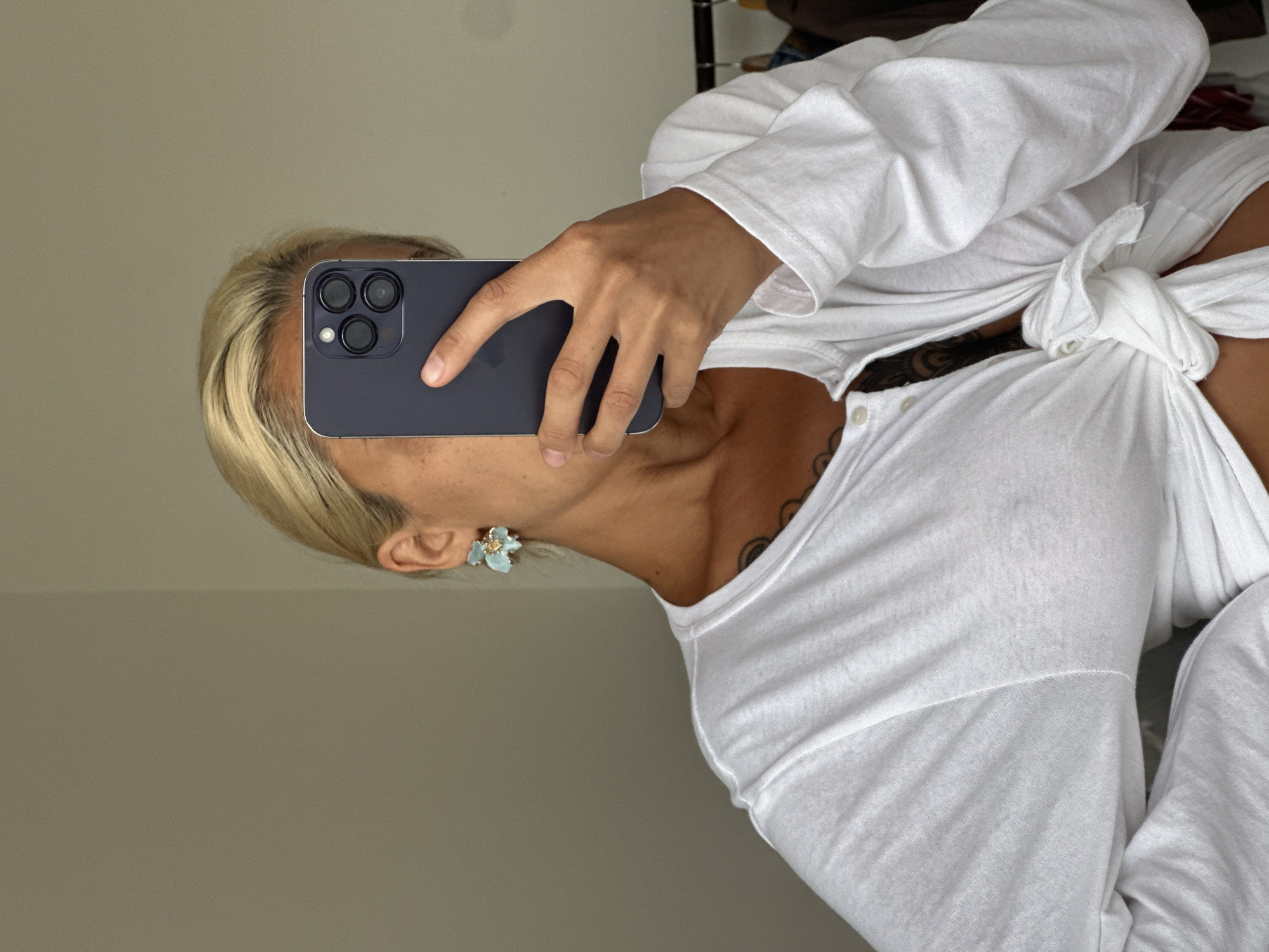 A person with blonde hair, wearing a white long-sleeved shirt tied at the waist and floral earrings, is taking a mirror selfie in Kuta Selatan, Indonesia. The background appears to be an indoor setting with a plain wall and a rack of clothes visible on the right. The person is holding a dark-colored smartphone, obscuring their face.