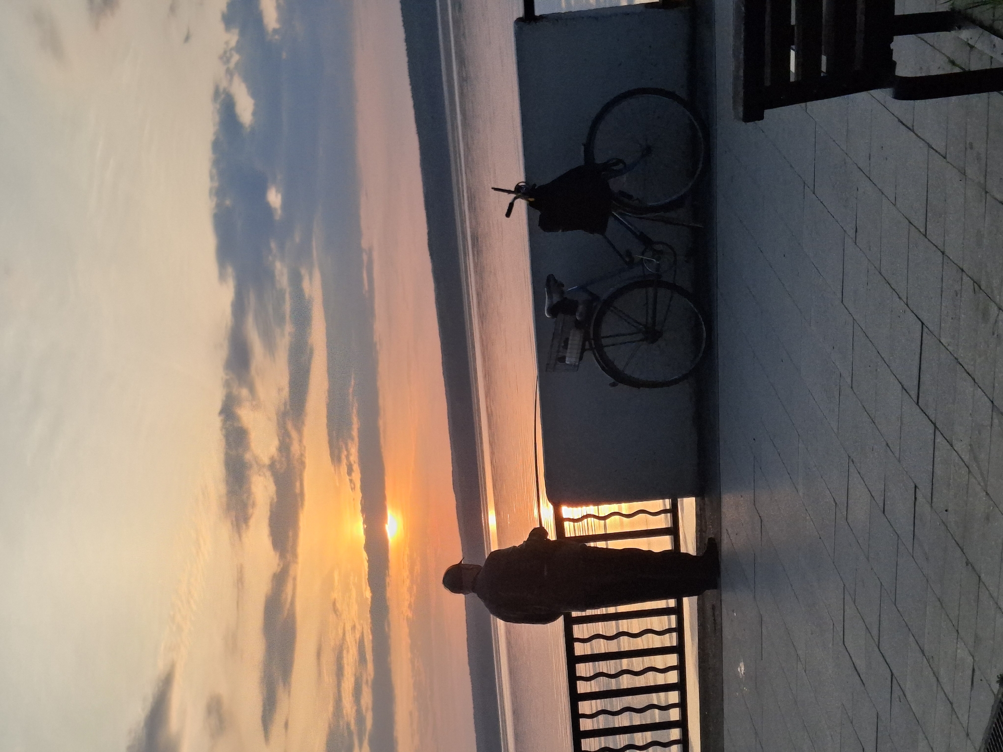 The image depicts a person standing on a tiled waterfront promenade in Chaikovsky, Russia, silhouetted against a sunset. The sunset illuminates the water, creating a bright reflection. A bicycle is parked against a concrete retaining wall, and a metal railing separates the walkway from the water. The scene appears to be taken during twilight or early evening.
