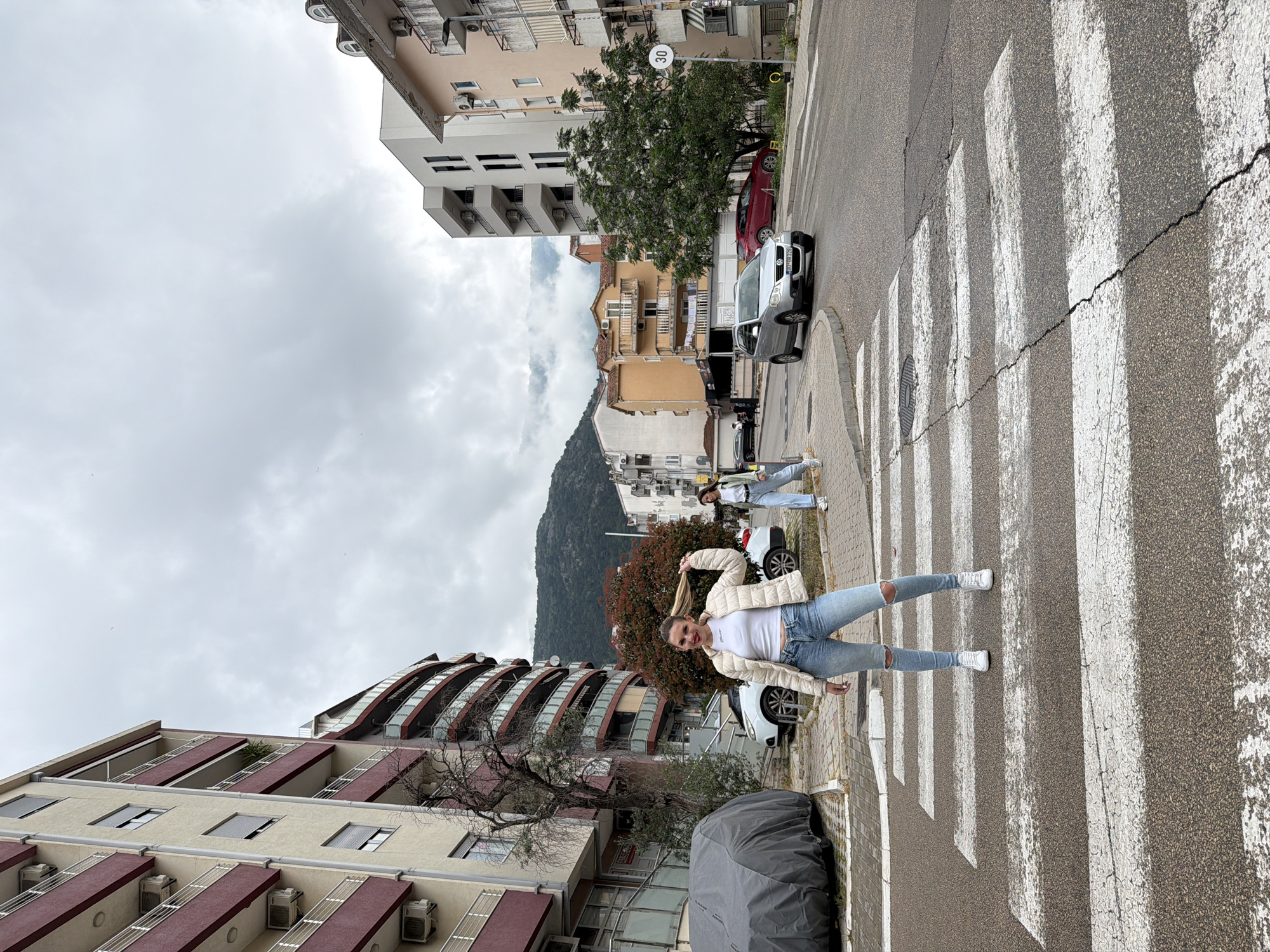 The image captures a street scene in Budva, Montenegro, notable for its architectural backdrop and the presence of pedestrians on a crosswalk.
The primary subject is a young woman standing on the crosswalk, striking a pose while holding her ponytail. She is clad in jeans, a white top, a light-colored jacket, and white sneakers. Another individual is visible in the background, further down the crosswalk, similarly dressed in casual attire.
The setting features a street with a pedestrian crosswalk. Buildings of varied architectural styles are on either side of the street. One building is beige with red trim and features balconies with air conditioning units. Another building is white with a modern, curved design. A mountain range is visible in the distance, adding depth to the scene. The sky is overcast. Several cars are parked alongside the road.